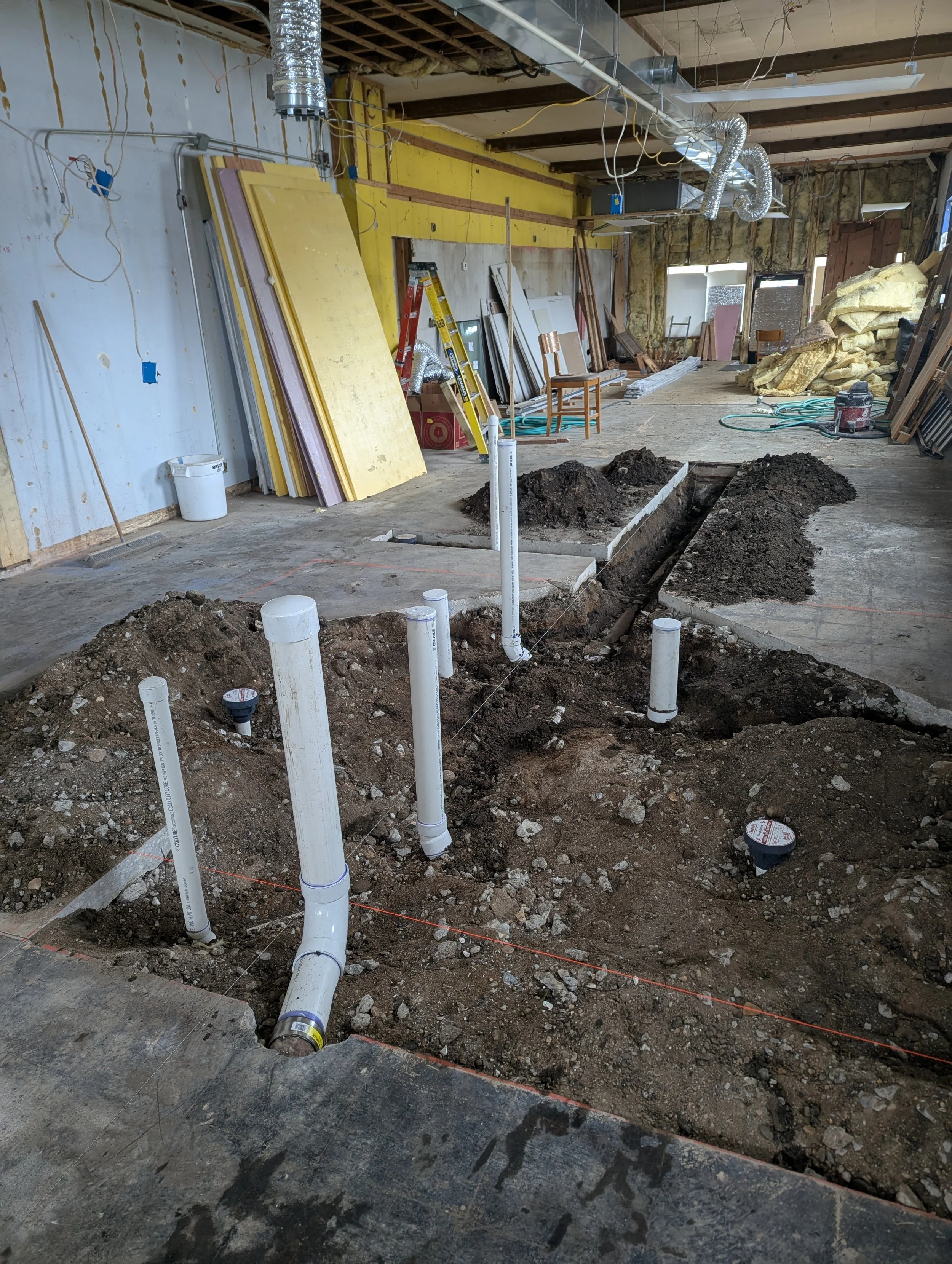 Interior of a building under construction with exposed pipes, electrical wiring, and construction materials scattered around. Dug trenches in the floor for plumbing installation.