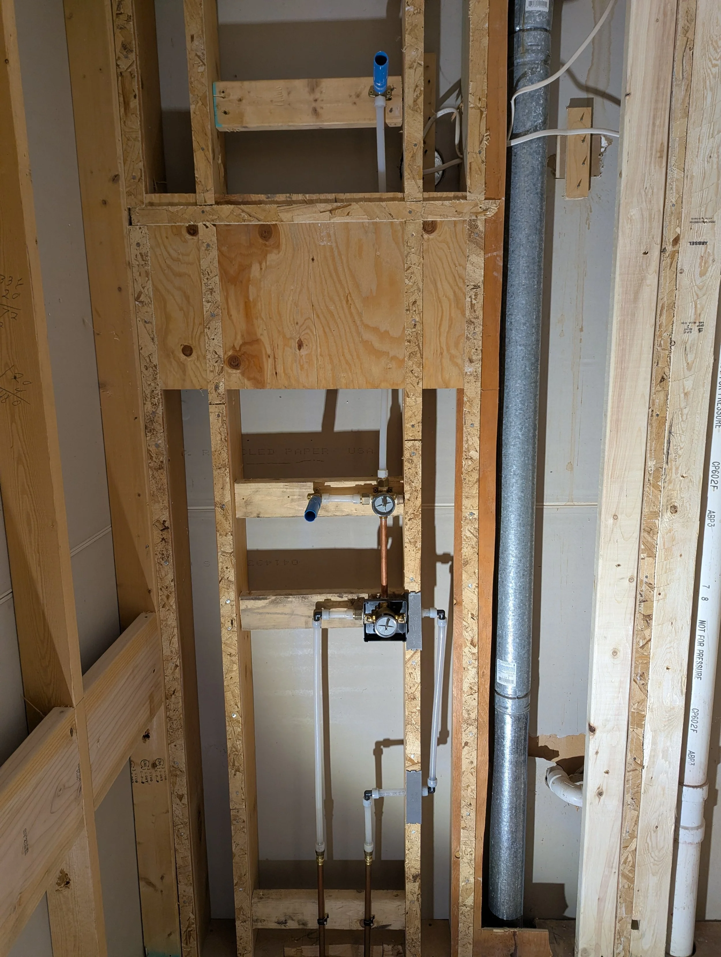 View of a plumbing installation within a wooden wall frame, showing pipes, valves, and a pressure gauge for a bathroom or kitchen fixture.