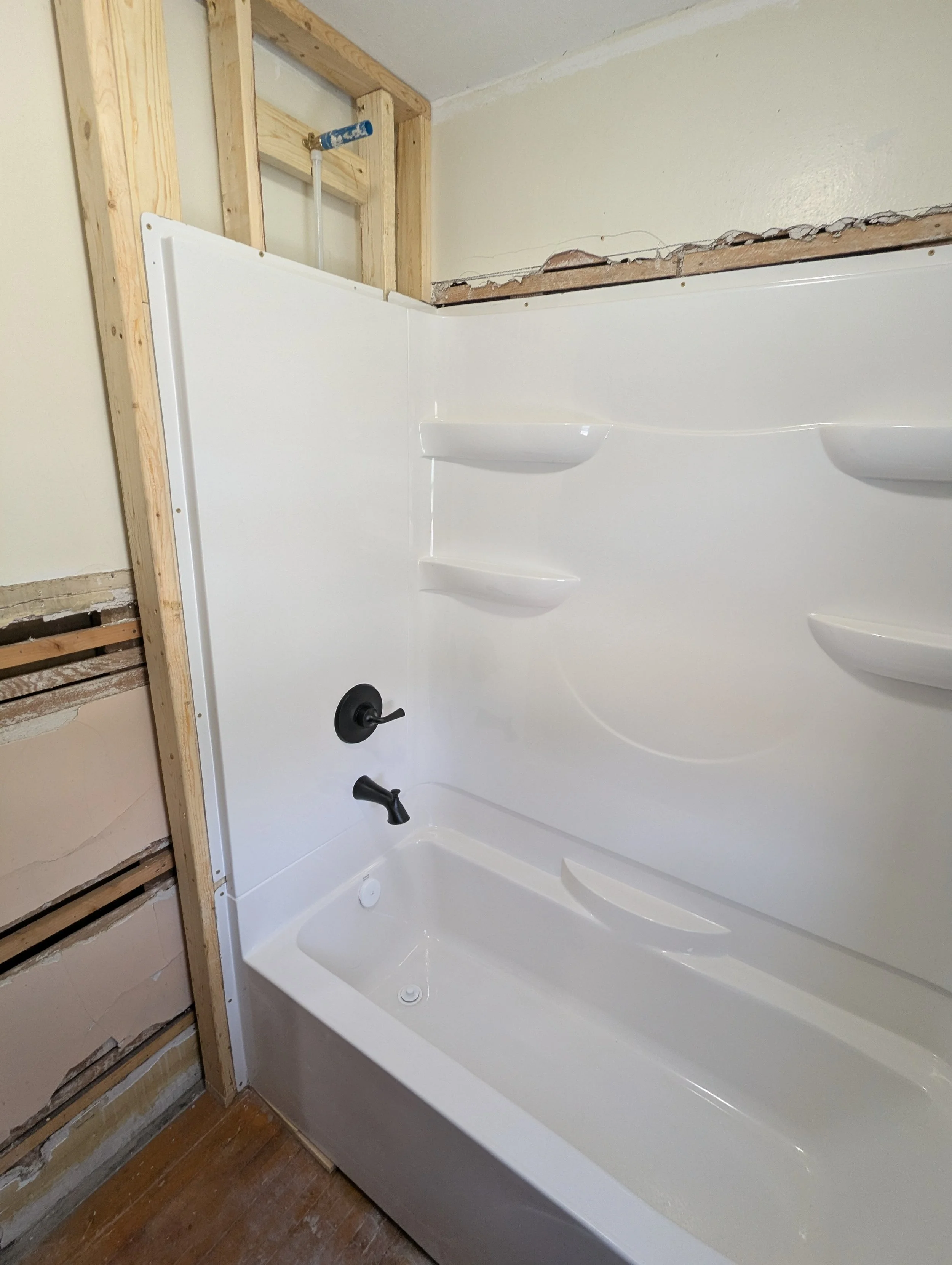 Newly installed white bathtub with black fixtures in a bathroom under renovation, with exposed wooden framing and wall in progress.