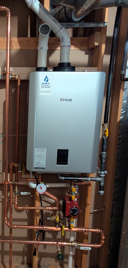 A Rinnai tankless water heater installed on a wooden wall in a plumbing setup, with copper pipes, a pressure gauge, and a red circulator pump.
