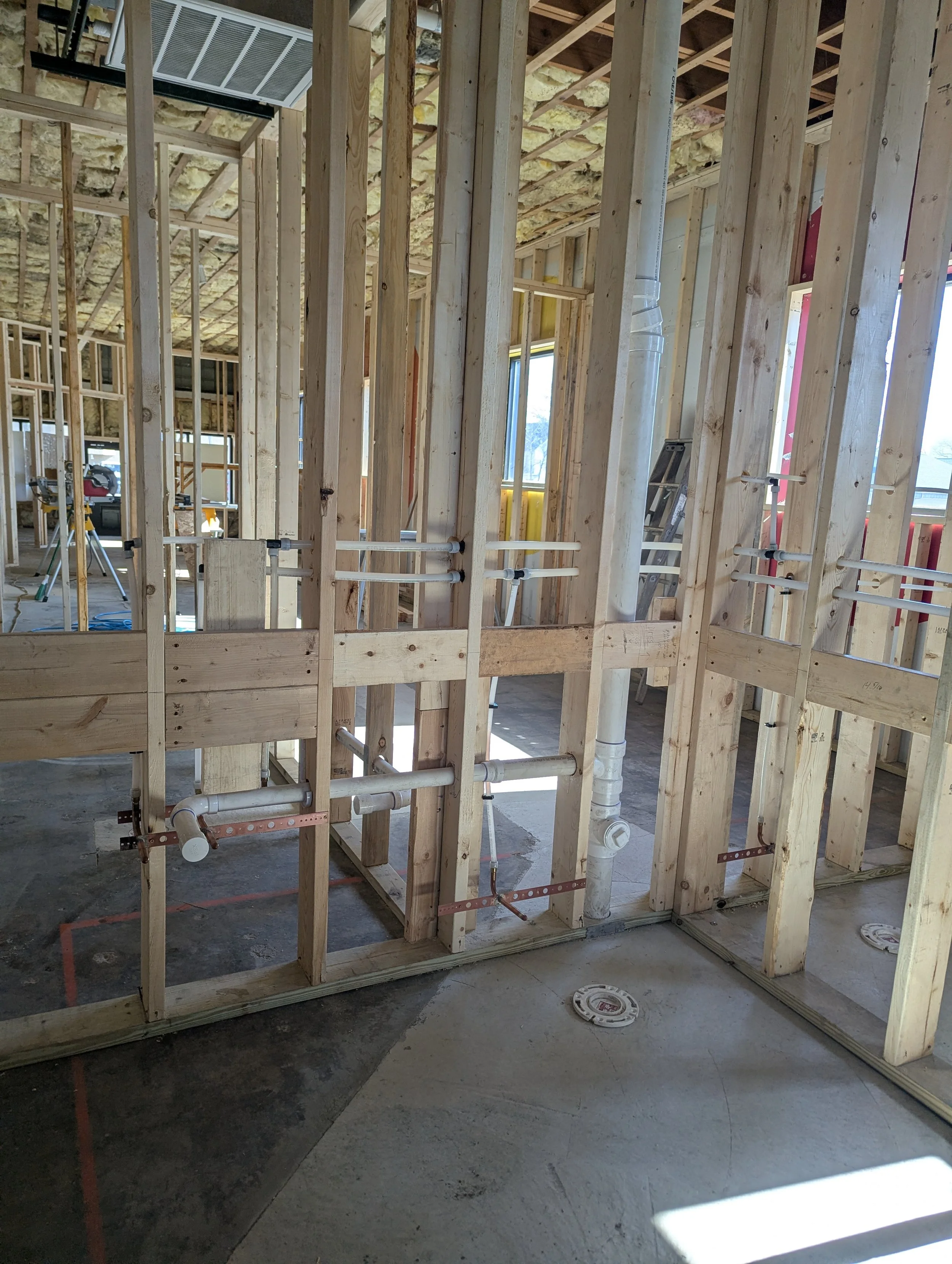 Interior view of a building under construction with exposed wooden framing, electrical wiring, and plumbing pipes.