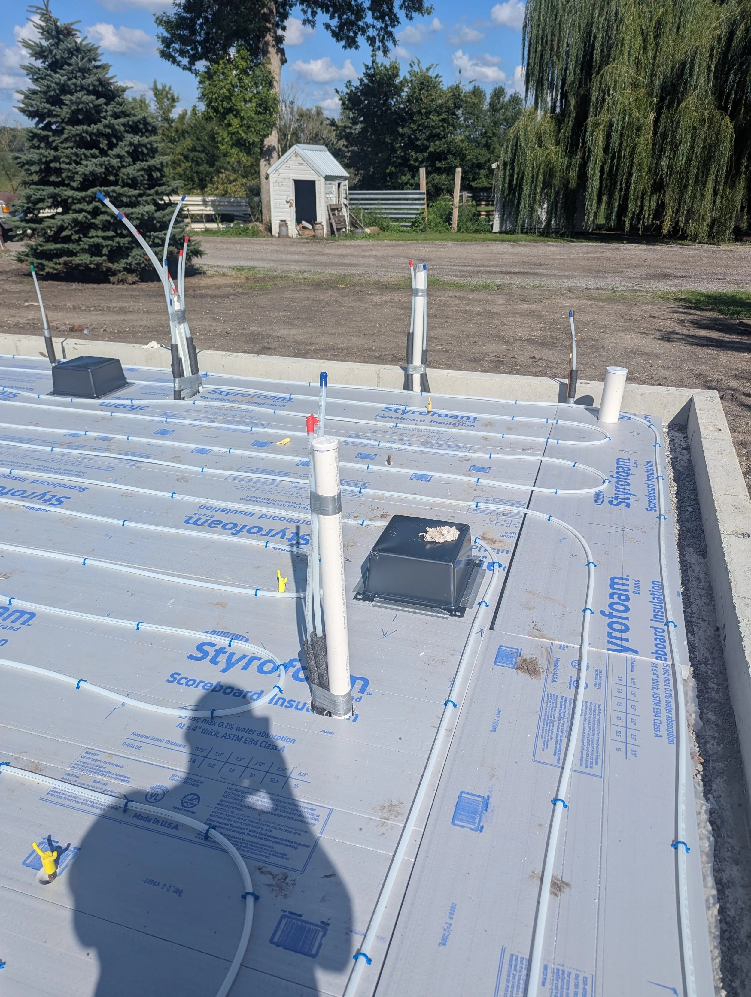 Construction site with plumbing pipes and Styrofoam insulation on a floor slab, with trees and a small shed in the background.