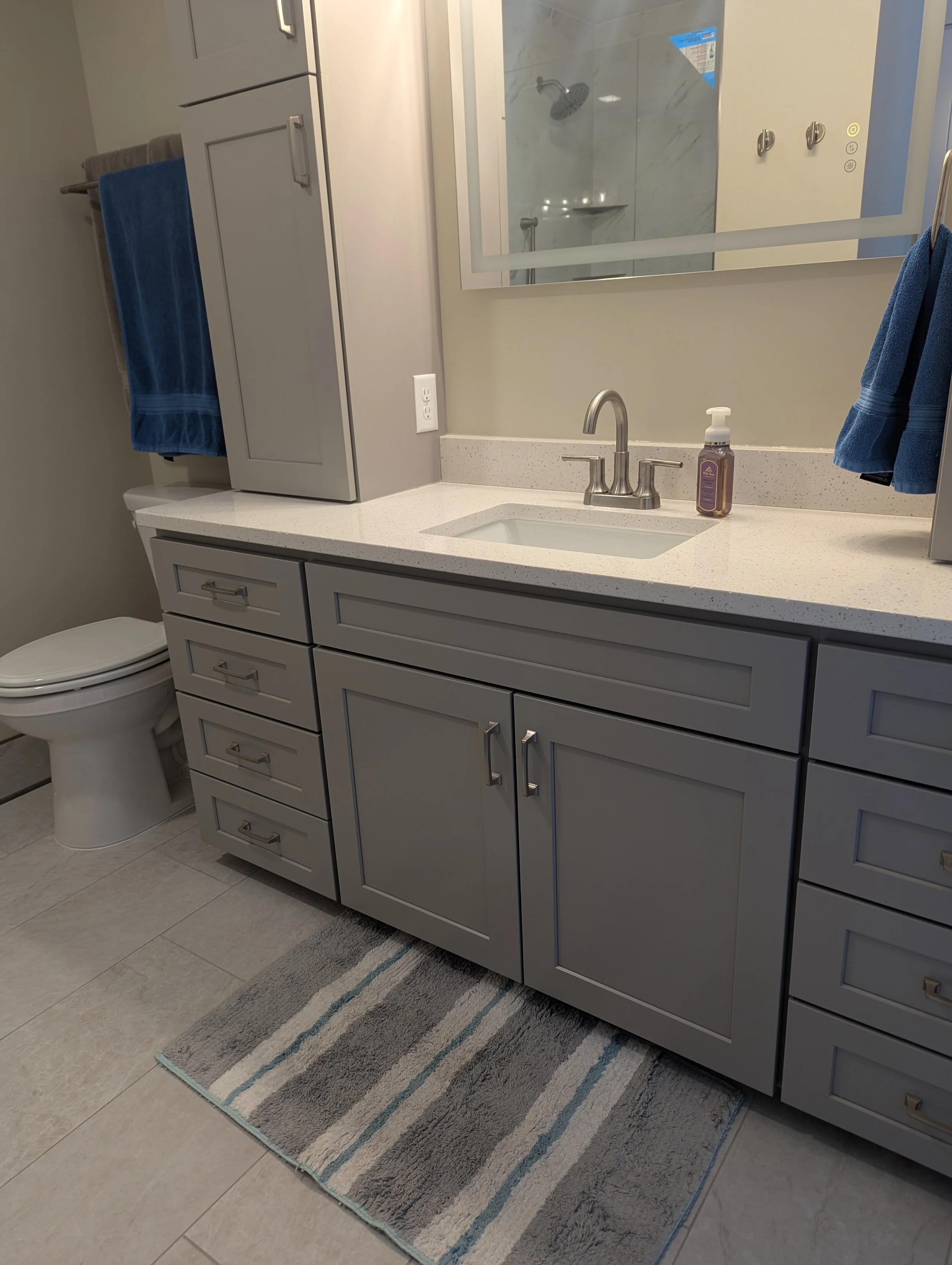 A bathroom vanity with a gray cabinet, a white countertop, a small sink, a chrome faucet, and a mirror. To the left, a toilet, and above it, a blue towel on a towel rack. A soap dispenser is on the countertop. There is also a gray and blue striped ru