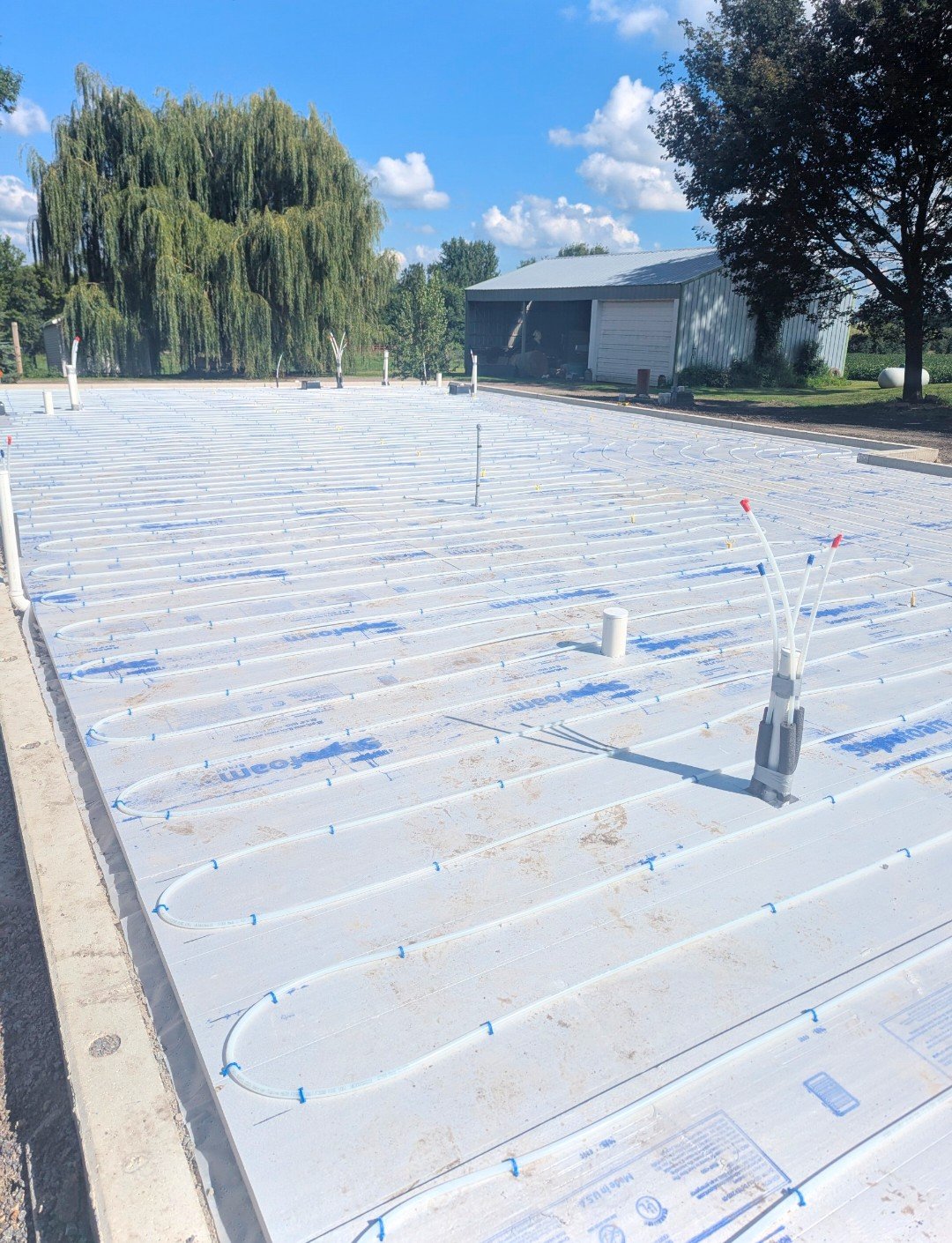 Undergoing installation of radiant floor heating system on a flat surface, with white tubing arranged in loops and pipes protruding from the setup, outdoor setting with a blue sky, trees, and a building in the background.