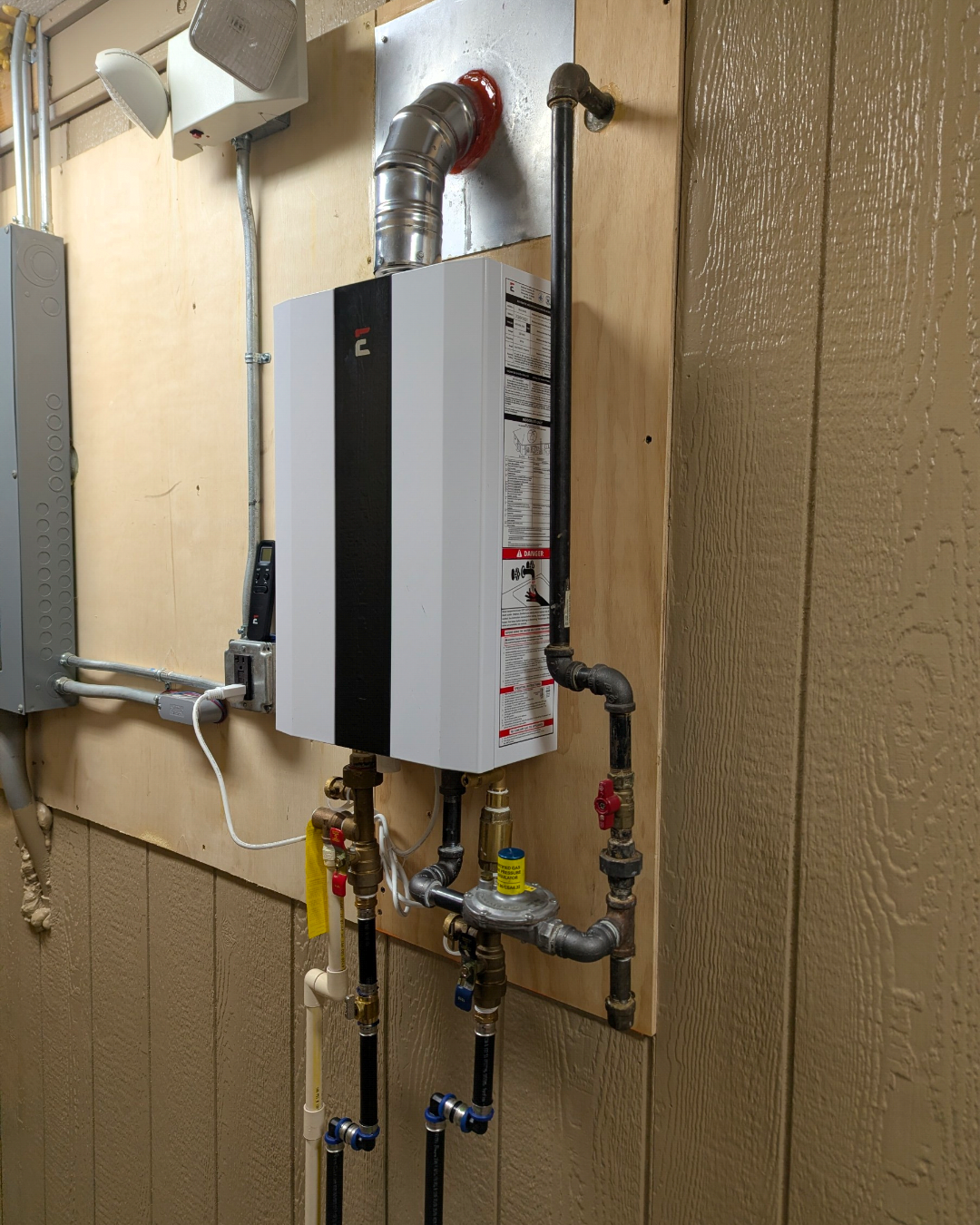 View of a water heater mounted on a wall with plumbing pipes, electrical wires, and a control panel nearby.
