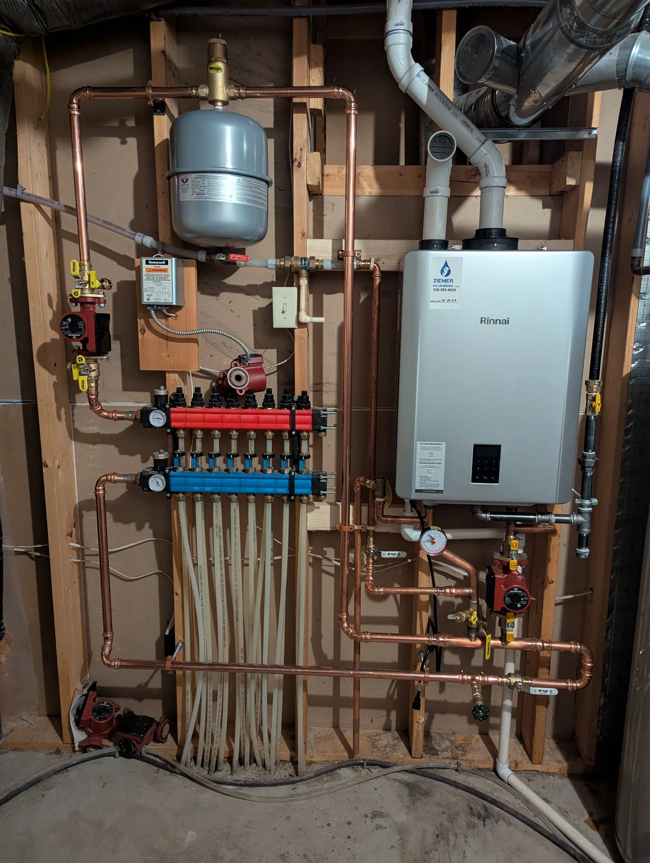 Indoor plumbing system with copper pipes, a water heater, a pressure regulator, and gauges mounted on a wooden wall frame.