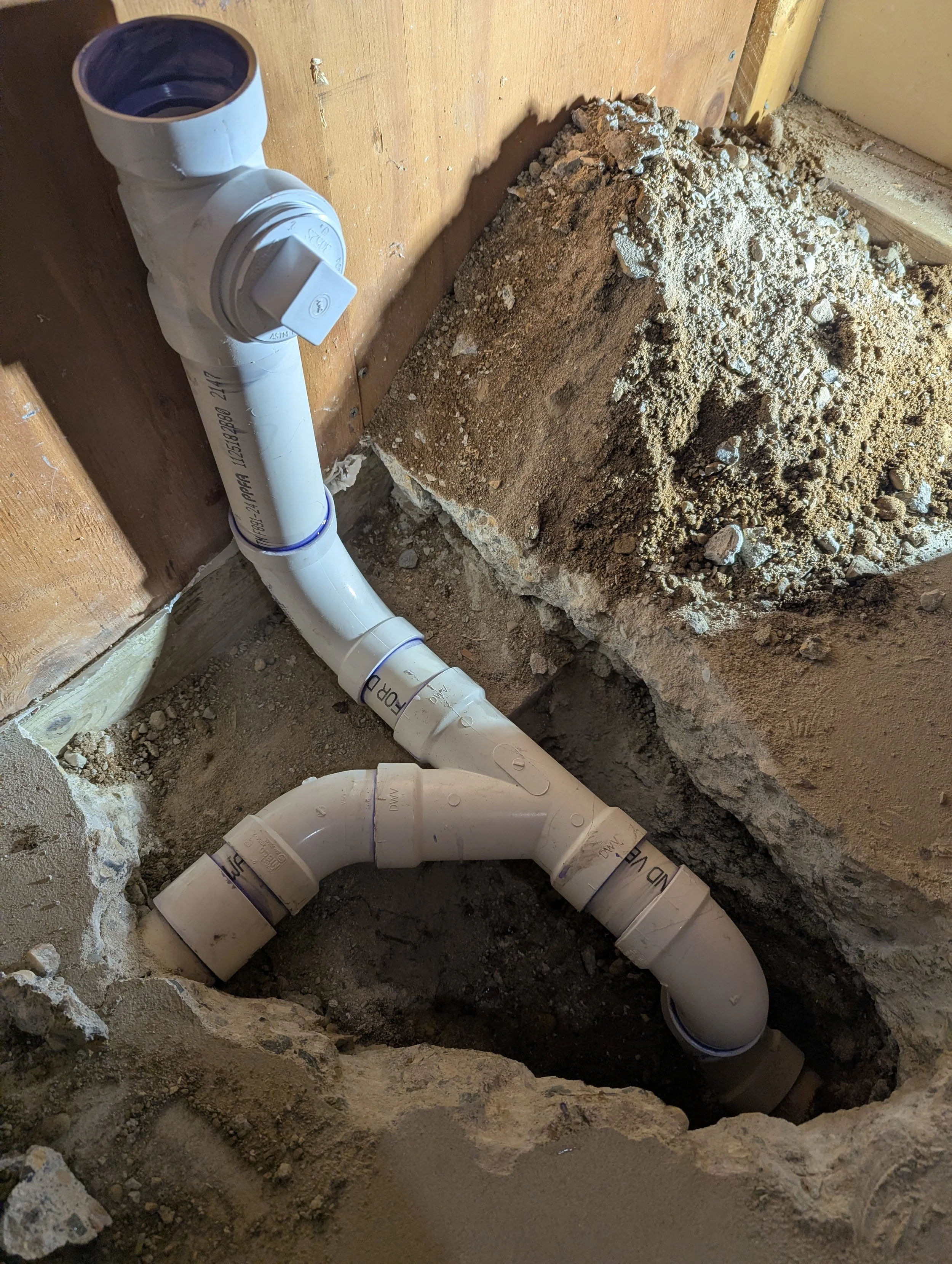 Close-up of PVC plumbing pipes and fittings in a construction or renovation site, with soil and debris around.