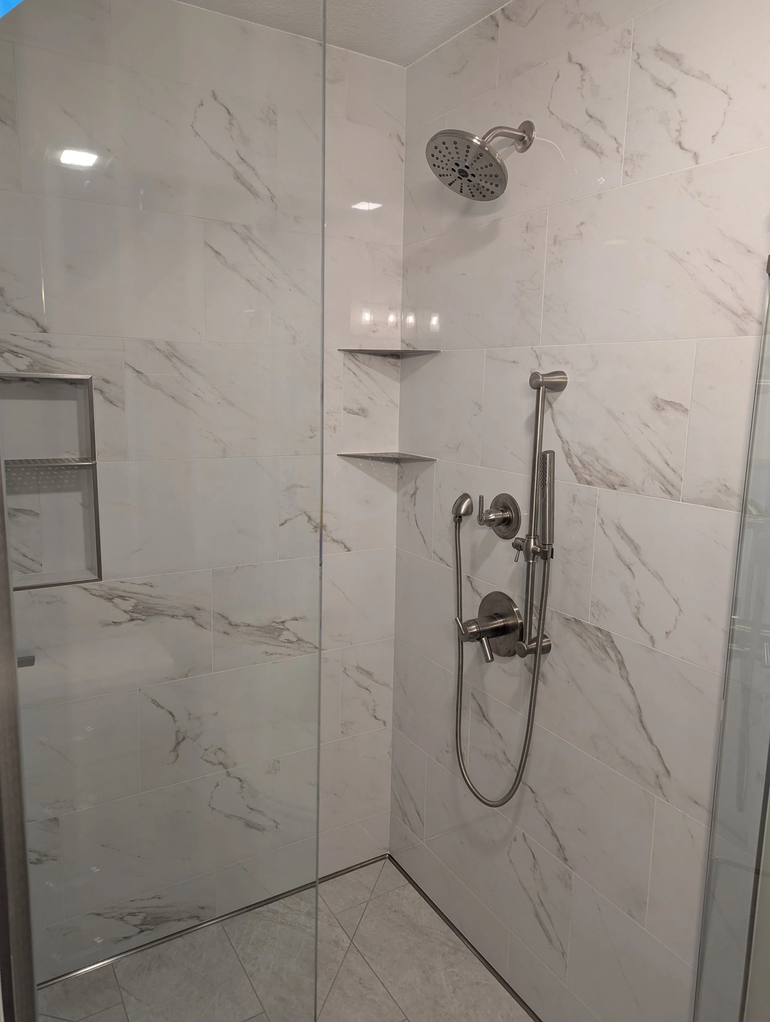 A modern shower with white marble tiles, a rain showerhead, a handheld showerhead, and built-in corner shelves.