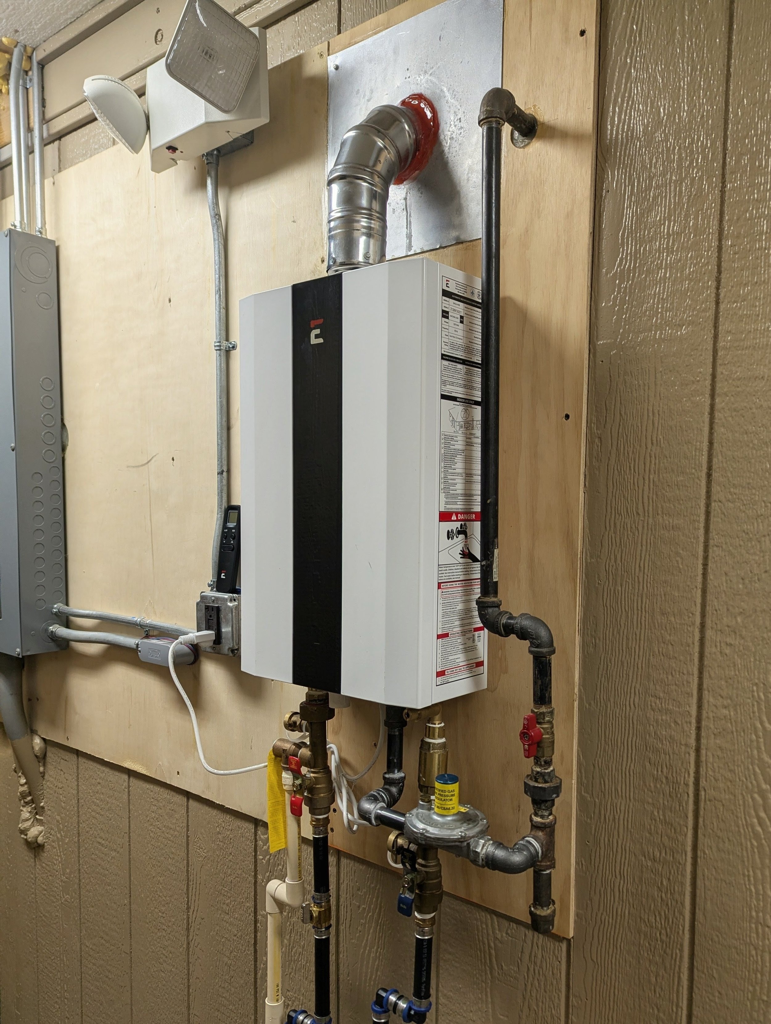 A wall-mounted tankless water heater with a vent pipe, various pipes, valves, and electrical components.