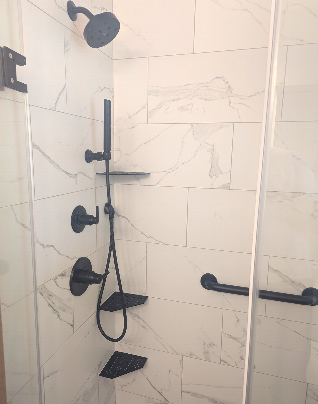 Shower with black fixtures, a handheld shower head, a wall-mounted shelf, and a grab bar in a white marble tiled shower enclosure.
