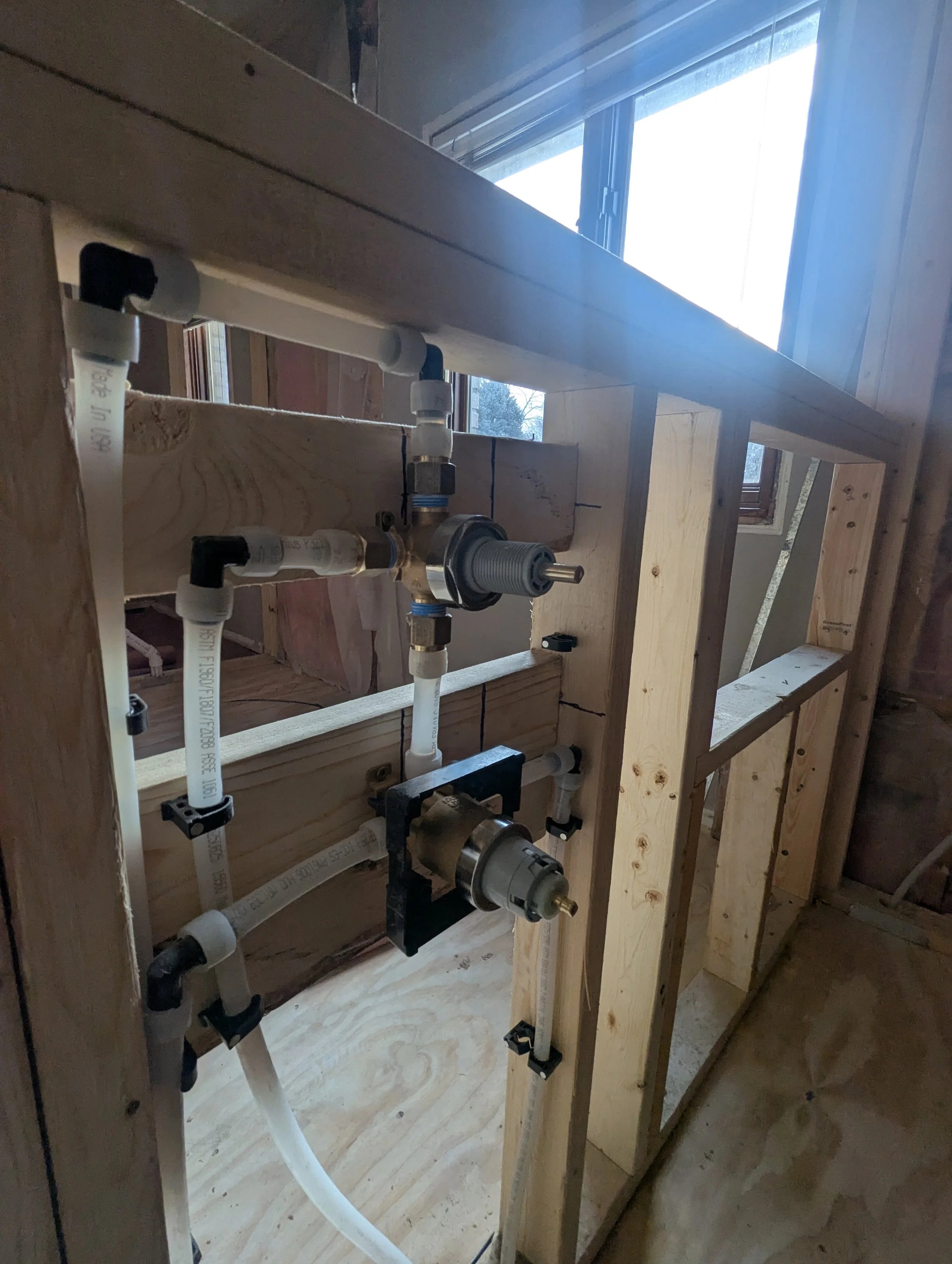 Partially constructed wall with plumbing and tubing installed, window in background, and framing for a wall in a building under construction.