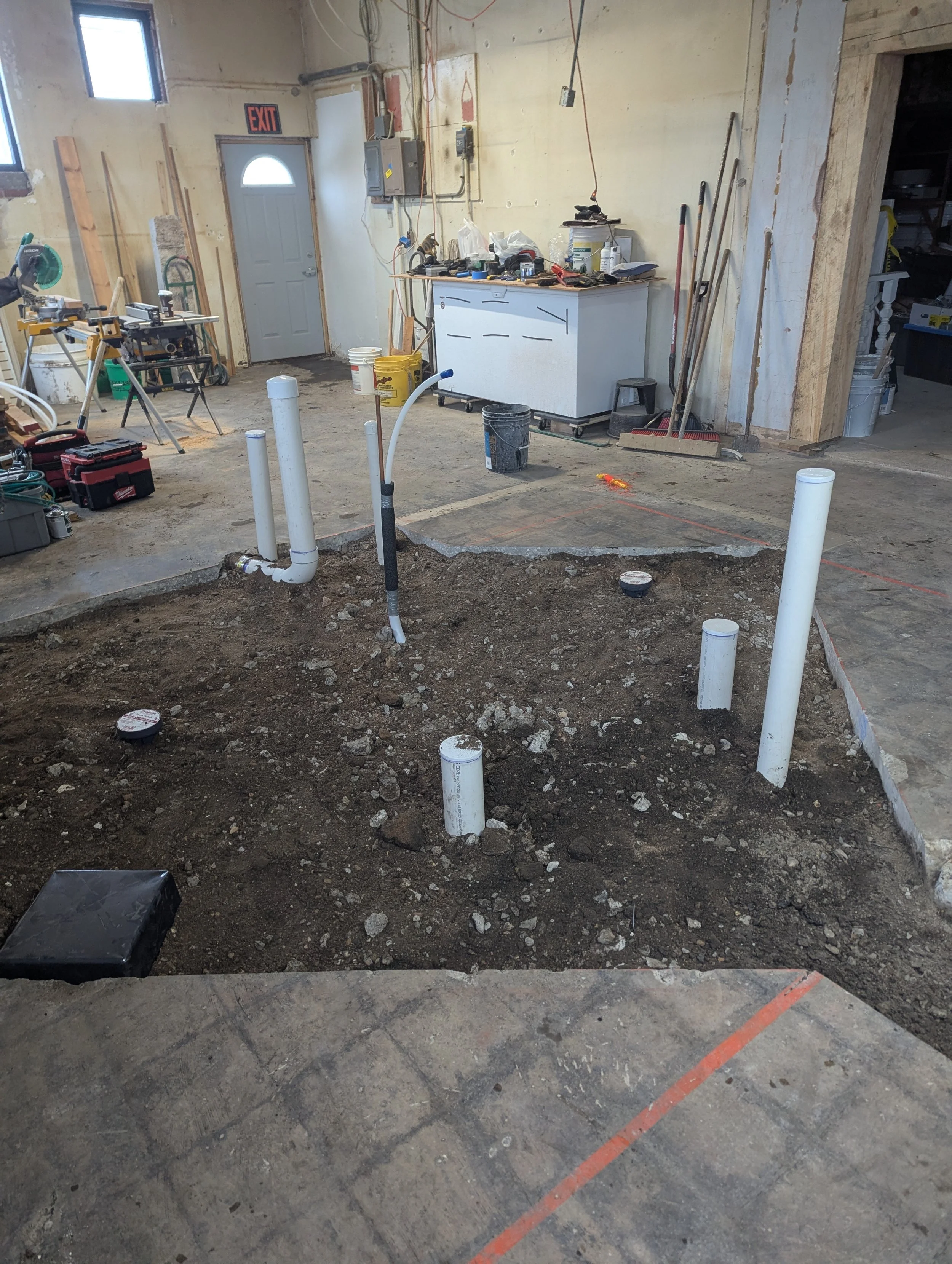 Indoor construction site with exposed concrete floor, PVC pipes set in the ground for plumbing, tools, buckets, and construction materials scattered around, some electrical wiring hanging from the ceiling, with a white cabinet and an exit door in the