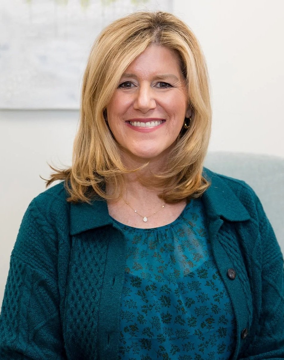 A woman with blonde hair smiling, wearing a teal patterned blouse and a matching teal cardigan, seated indoors with a white wall and a light-colored painting in the background.