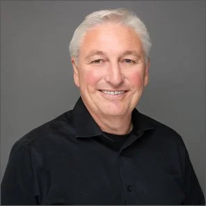 A smiling elderly man with white hair, wearing a black button-up shirt, against a plain gray background.