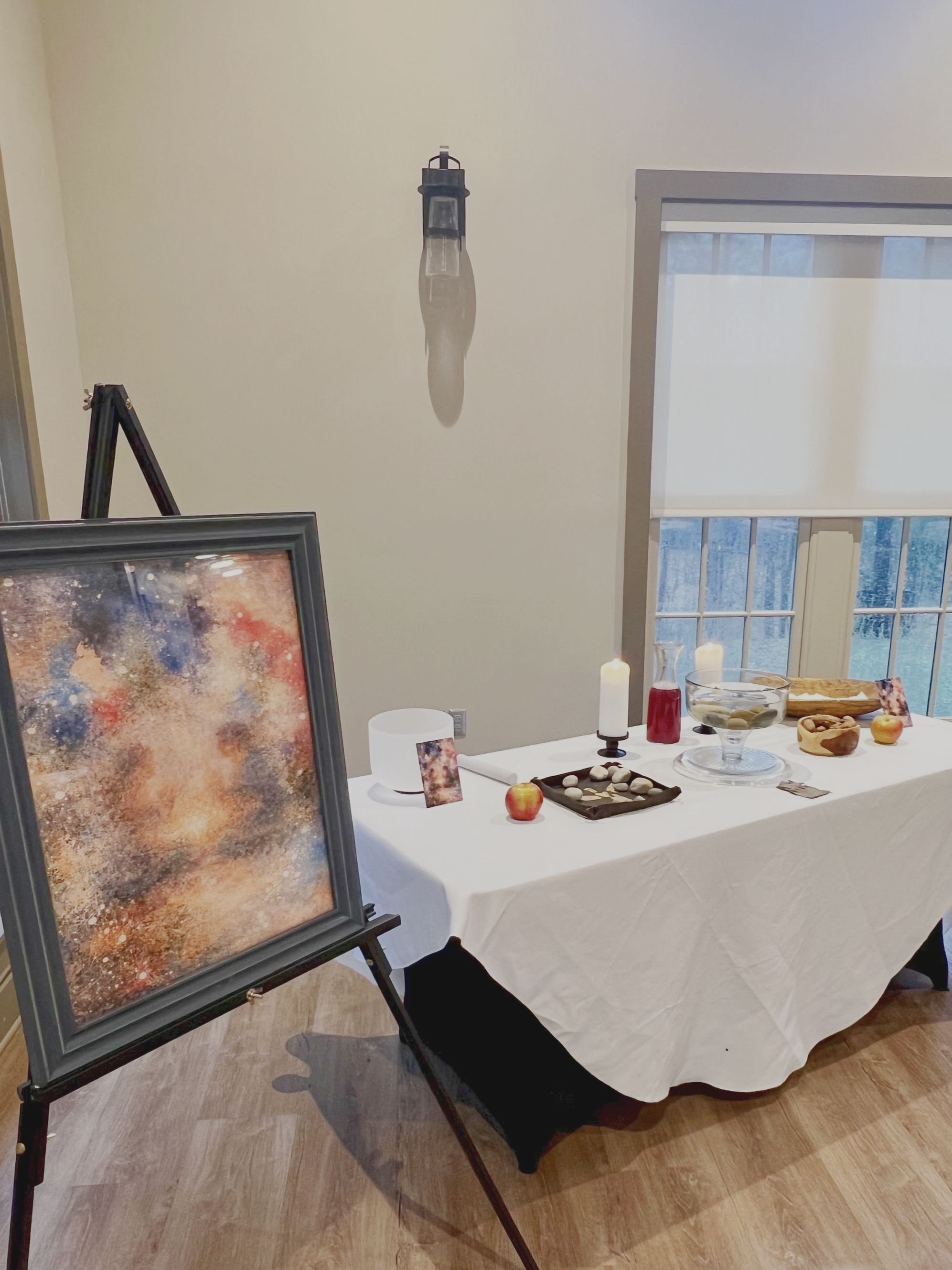 An art exhibit setup with a table draped in a white cloth, displaying various objects including candles, apples, and art pieces. A framed abstract painting on an easel and a light fixture hanging on the wall are also visible.