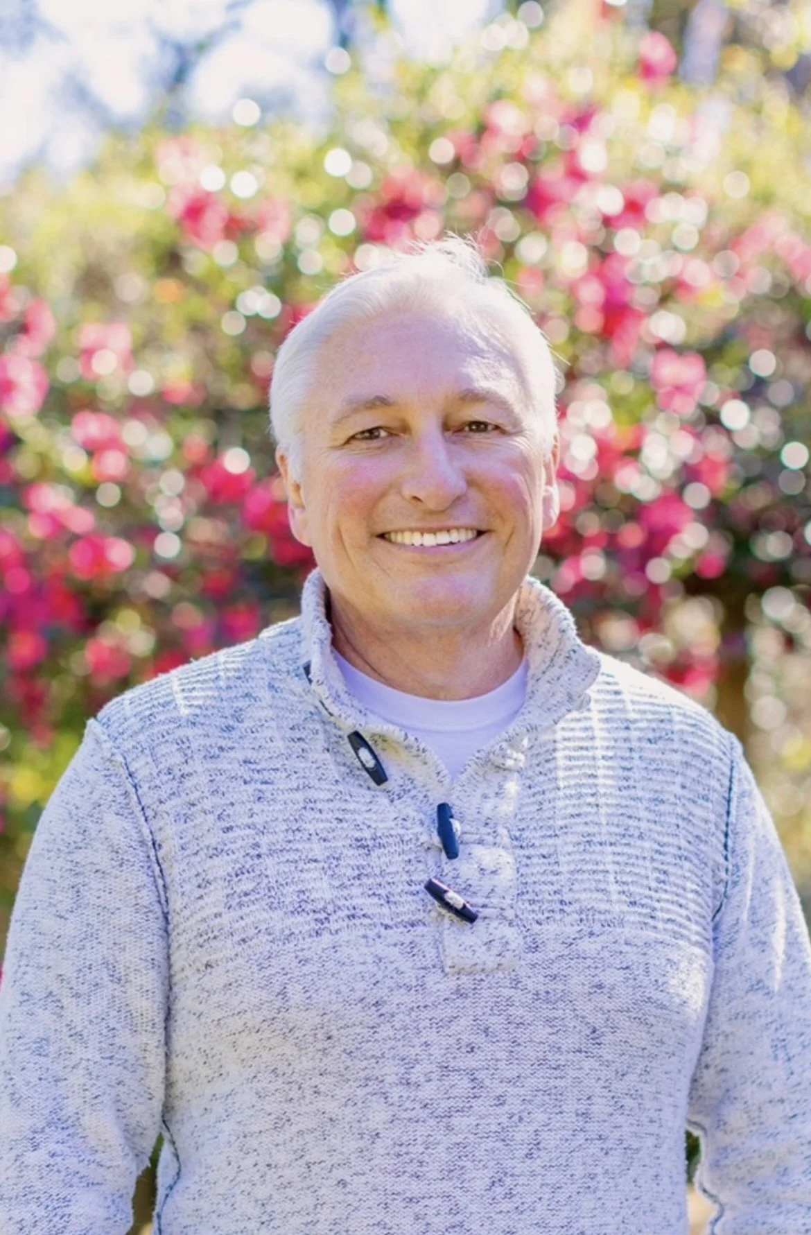 A smiling man with white hair, wearing a light gray quarter-zip sweater, standing outdoors in front of a colorful, blurred background of pink and green flowers and foliage.