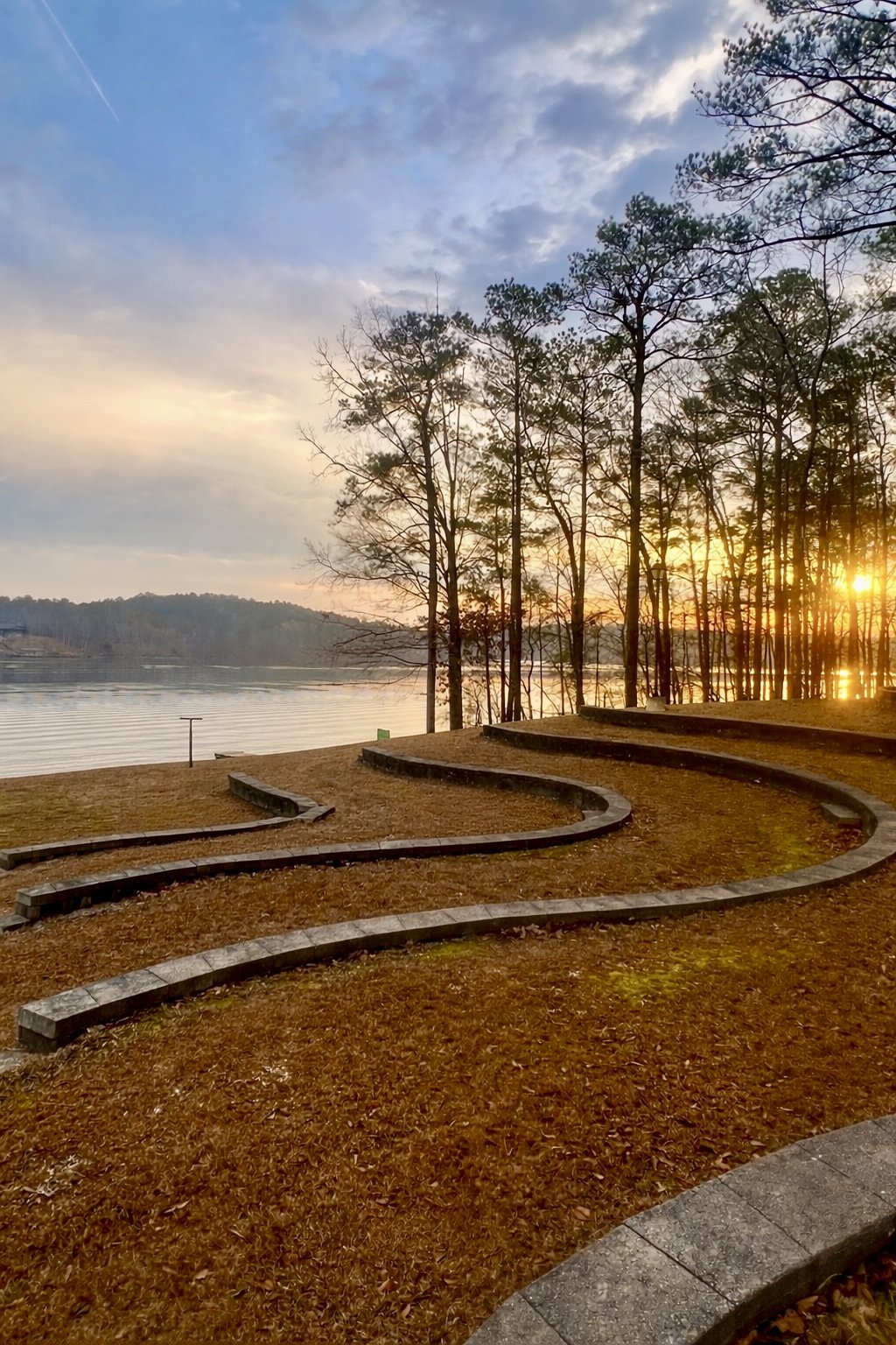 Sunset over a lake with leafless trees and a curved concrete path in the foreground.