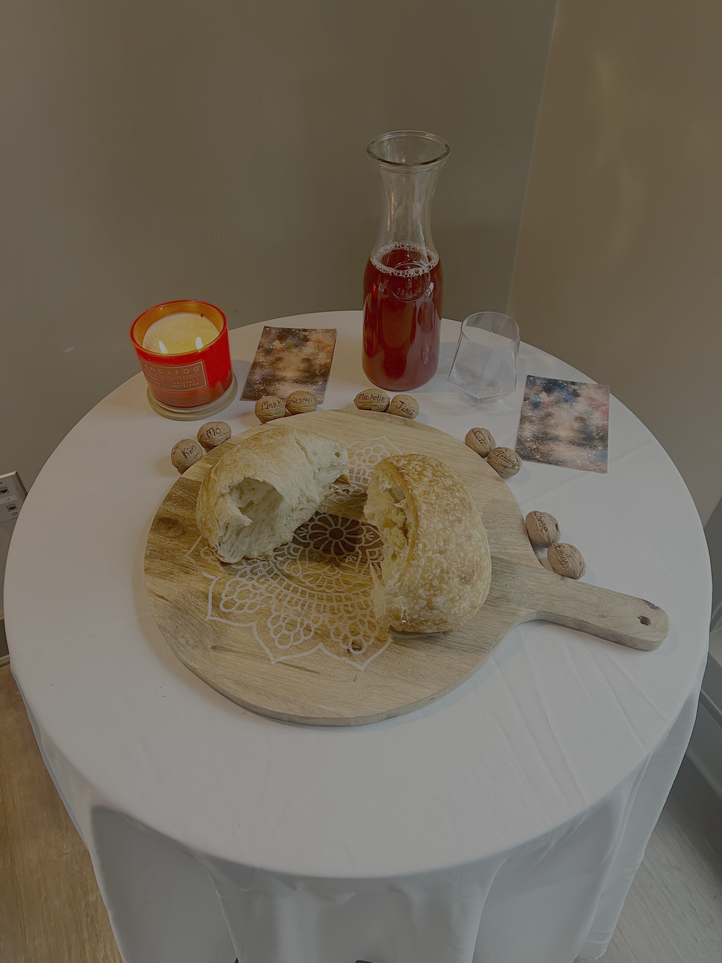 Bread on a wooden cutting board on a white table, with a bottle of red beverage, a glass, a red candle, and small stones with names written on them nearby.