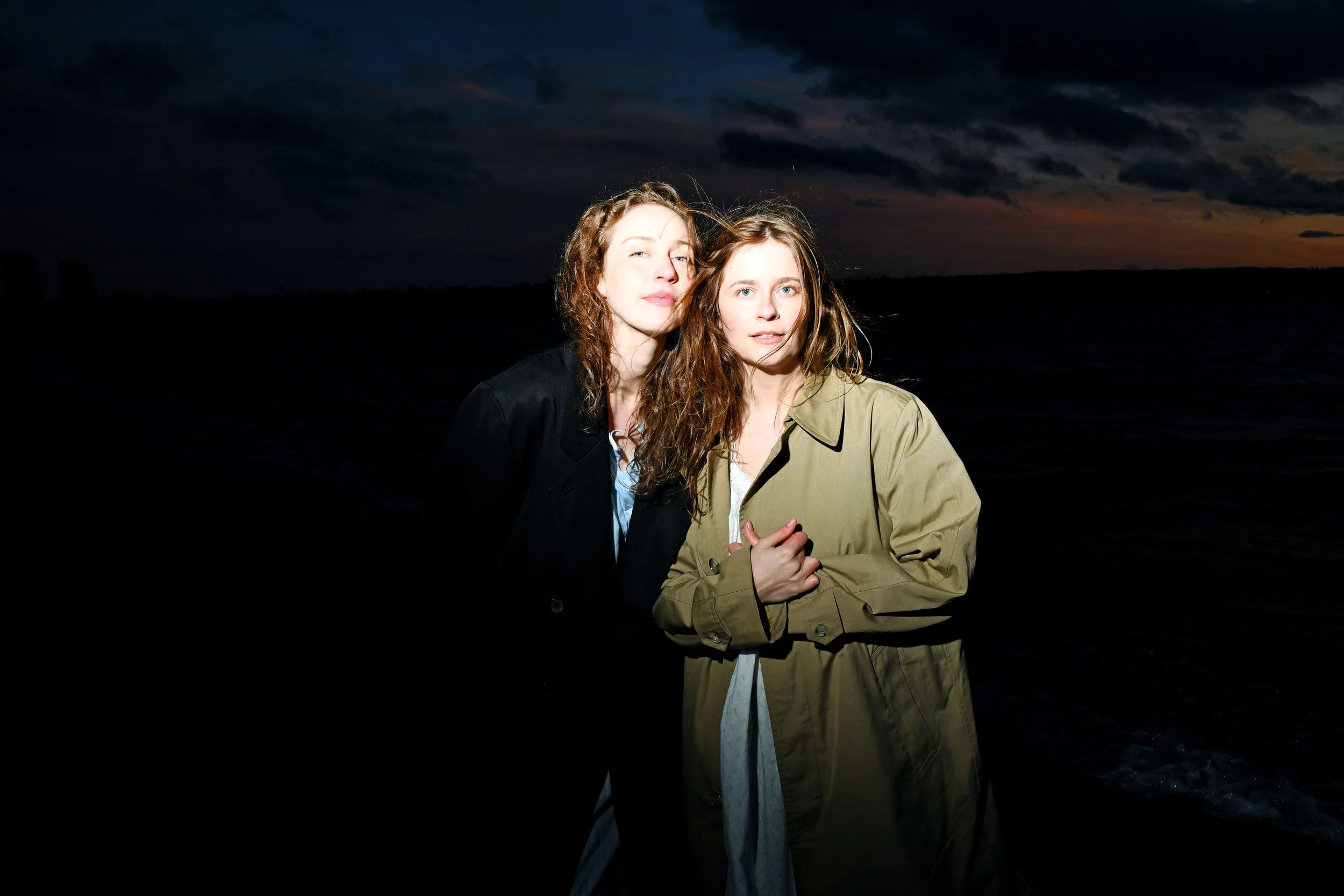 Two women with long, wavy hair standing close together outdoors at dusk, with a cloudy sky and sunset in the background.