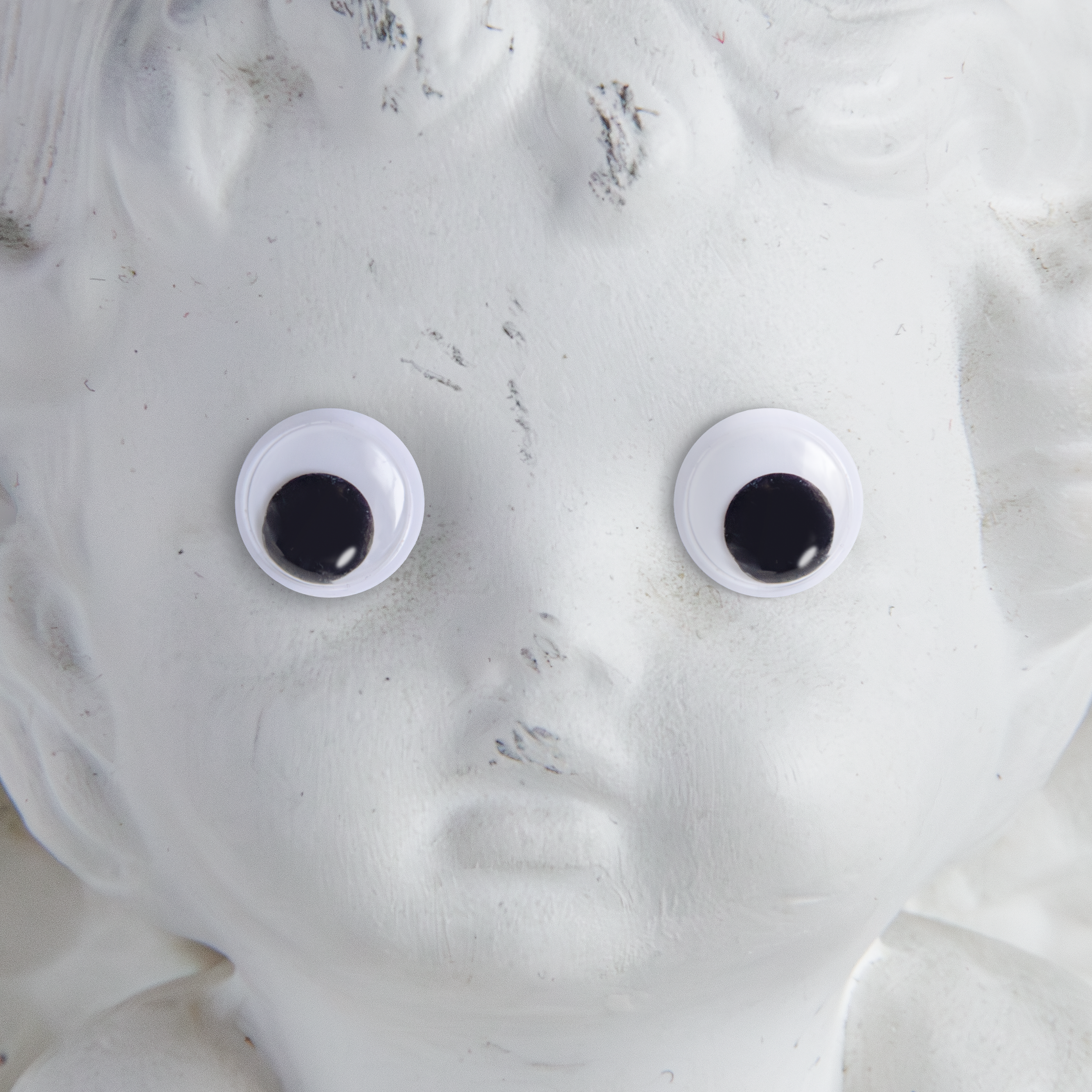 Close-up view of a cherub sculpture face with googly eyes and some dirt smudges