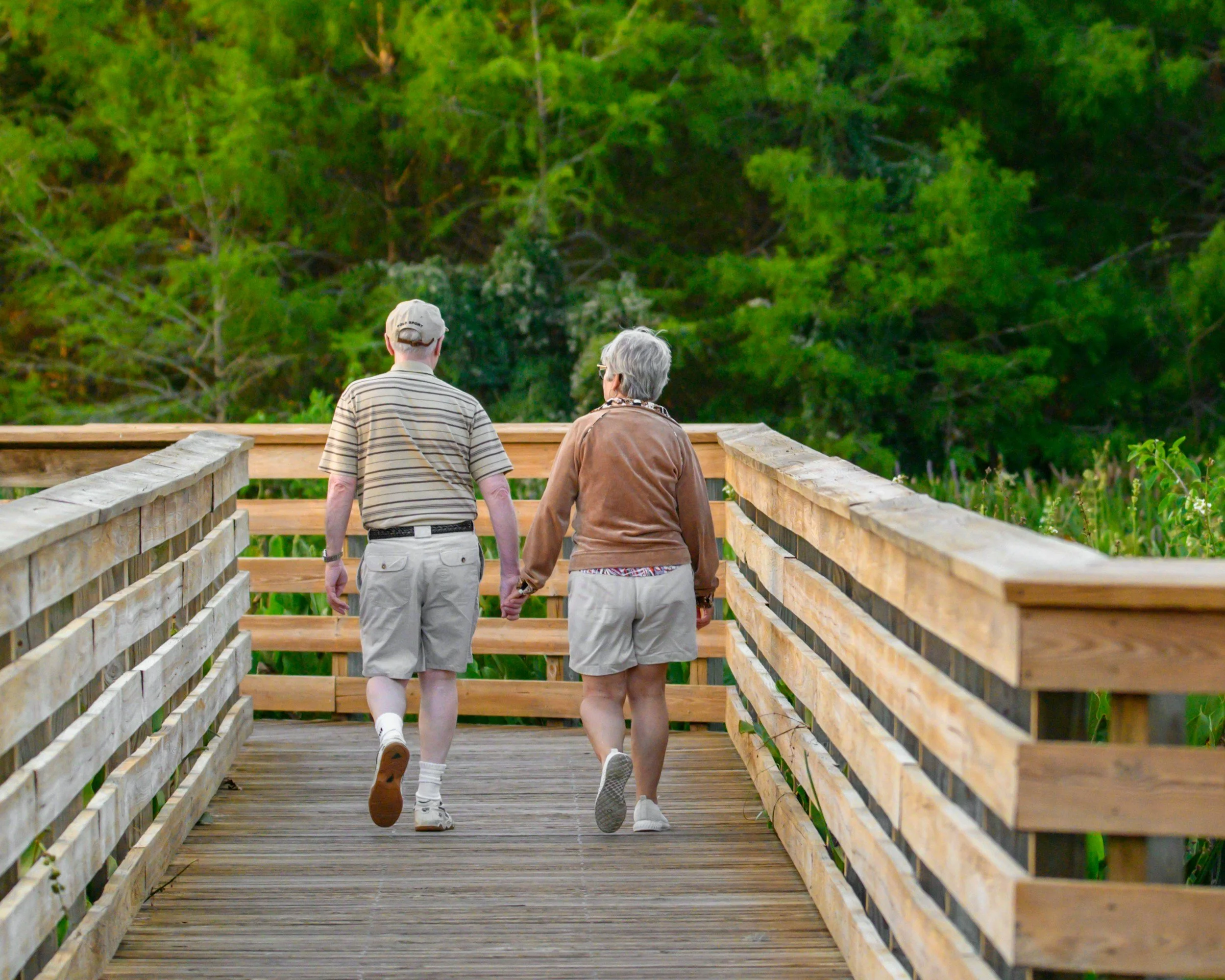 An elderly couple walking hand in hand on a wooden bridge surrounded by green trees.