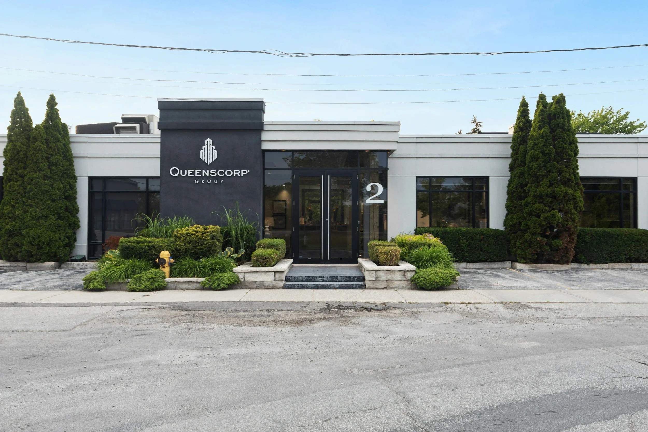 Front view of a modern office building with a sign that says 'QUEENSCORP GROUP' next to a glass entrance door labeled with the number 2, surrounded by well-kept bushes and trees.