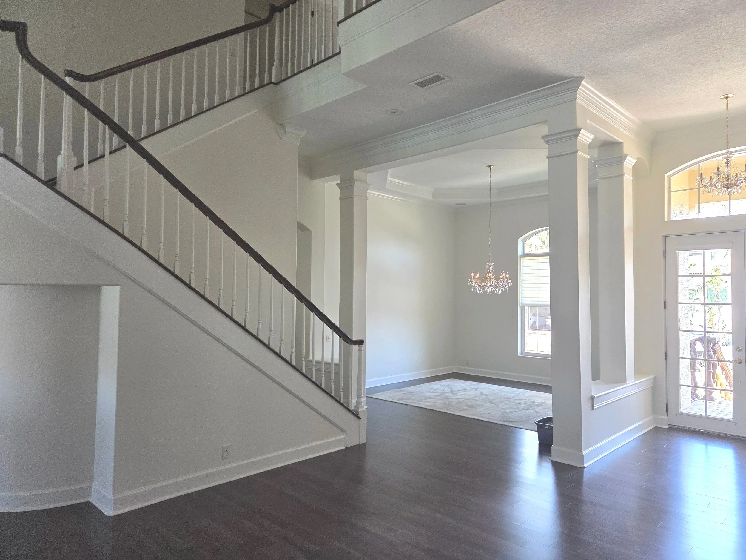 Interior view of a two-story house with white walls, dark hardwood flooring, and large windows. A staircase with white banisters is visible on the left. Elegant chandeliers hang from the ceiling, and decorative columns frame the entryway to another room.