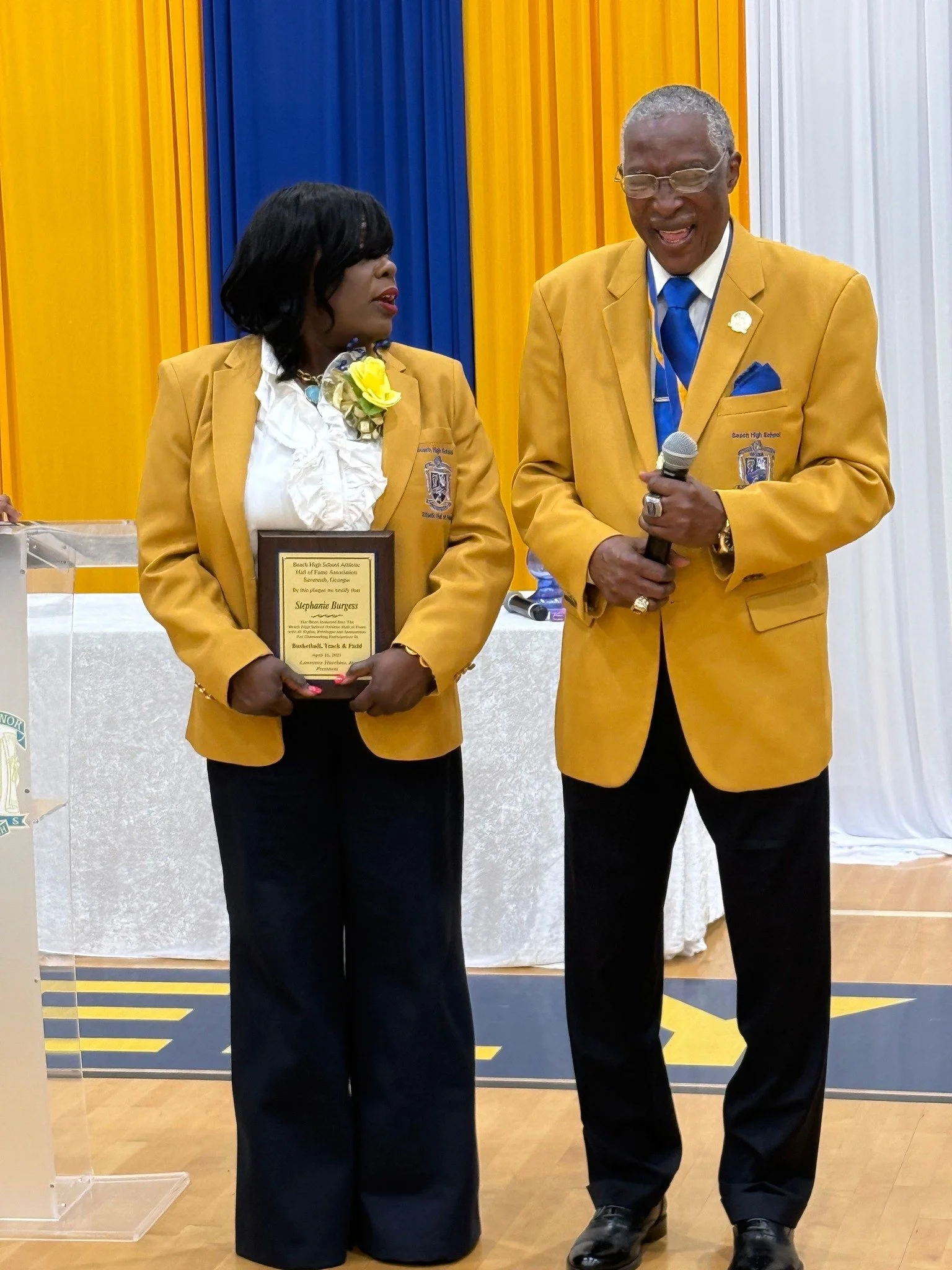 A woman and man wearing matching yellow blazers at an award ceremony. The woman is holding a plaque and wearing a white blouse with a yellow flower corsage, and the man is holding a microphone. The background features yellow, blue, and white curtains.
