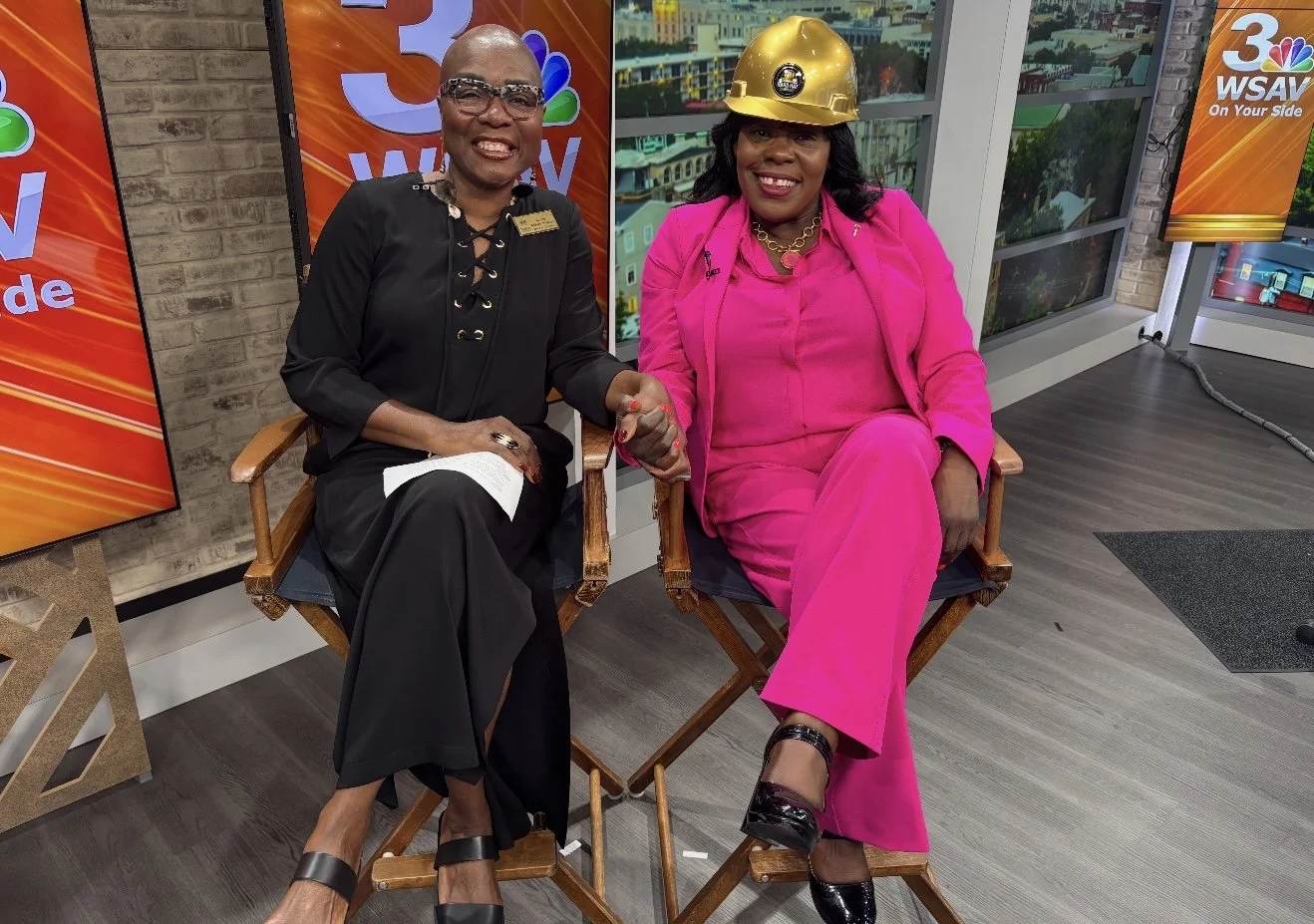 Two women sitting on director's chairs in a news studio, one wearing a black outfit and glasses, holding hands with the other woman in a bright pink suit and gold construction helmet, both smiling at the camera.