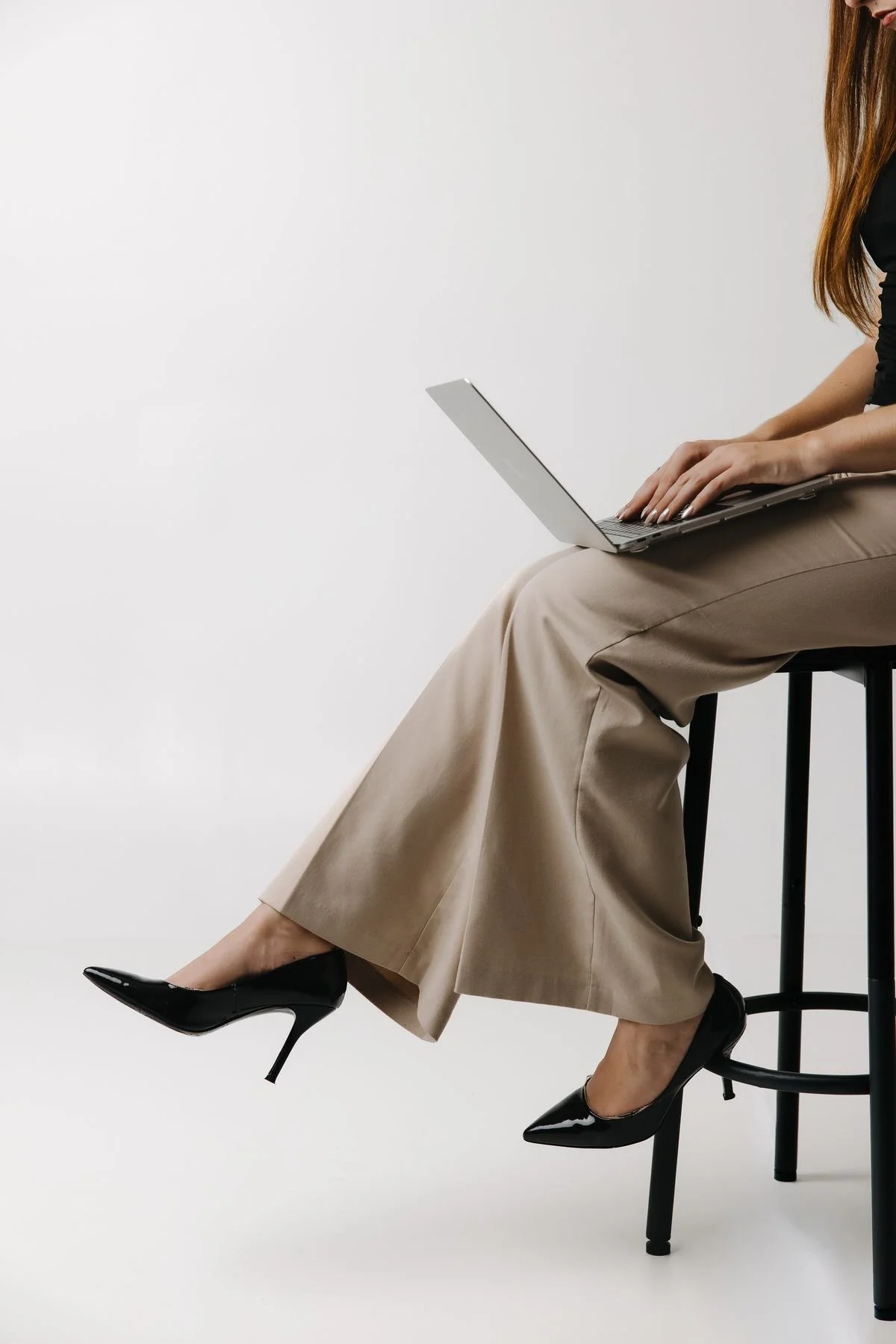 A woman sitting on a high black stool, wearing black high heels, beige wide-leg pants, a black top, and typing on a silver laptop against a plain white background.