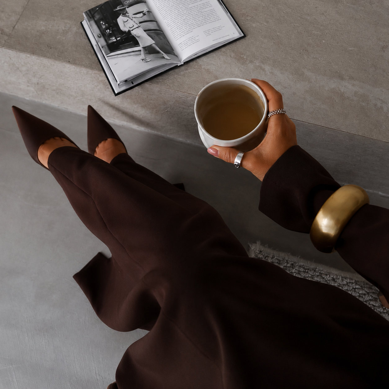 Sociél Muet Atelier Home Page - Cheri dressed in brown pants, brown pointed-toe shoes, holding a cup of coffee with a book open - Payroll Risk & Operations Consultant