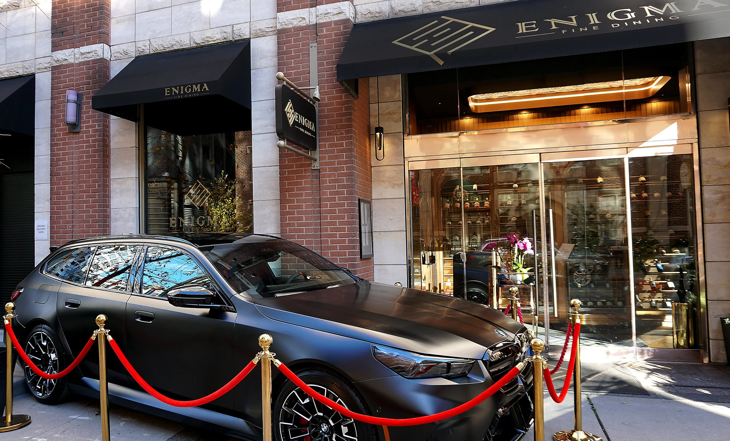 A sleek, matte black BMW car parked outside a high-end building with a jewelry store. The car is roped off with red velvet ropes and gold stanchions. The building's exterior features brick and marble, with black awnings and gold lettering for a fine 