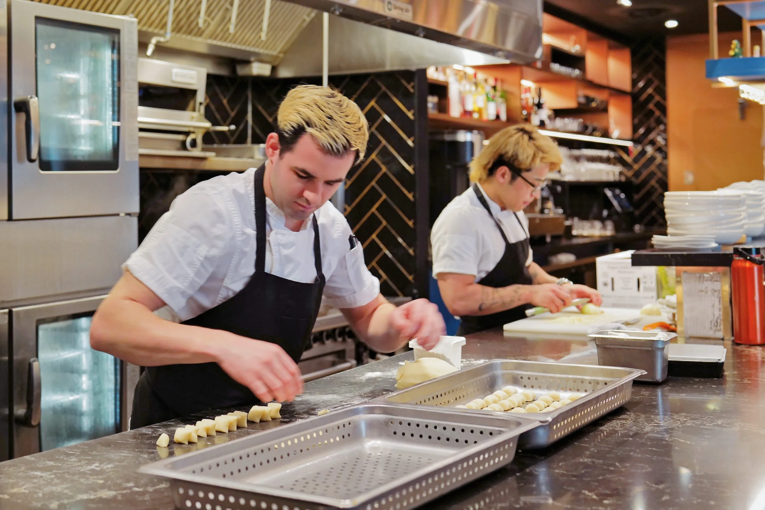 Two chefs in white shirts and black aprons preparing food in a commercial kitchen, working at a stainless steel counter with trays and chopping onions.