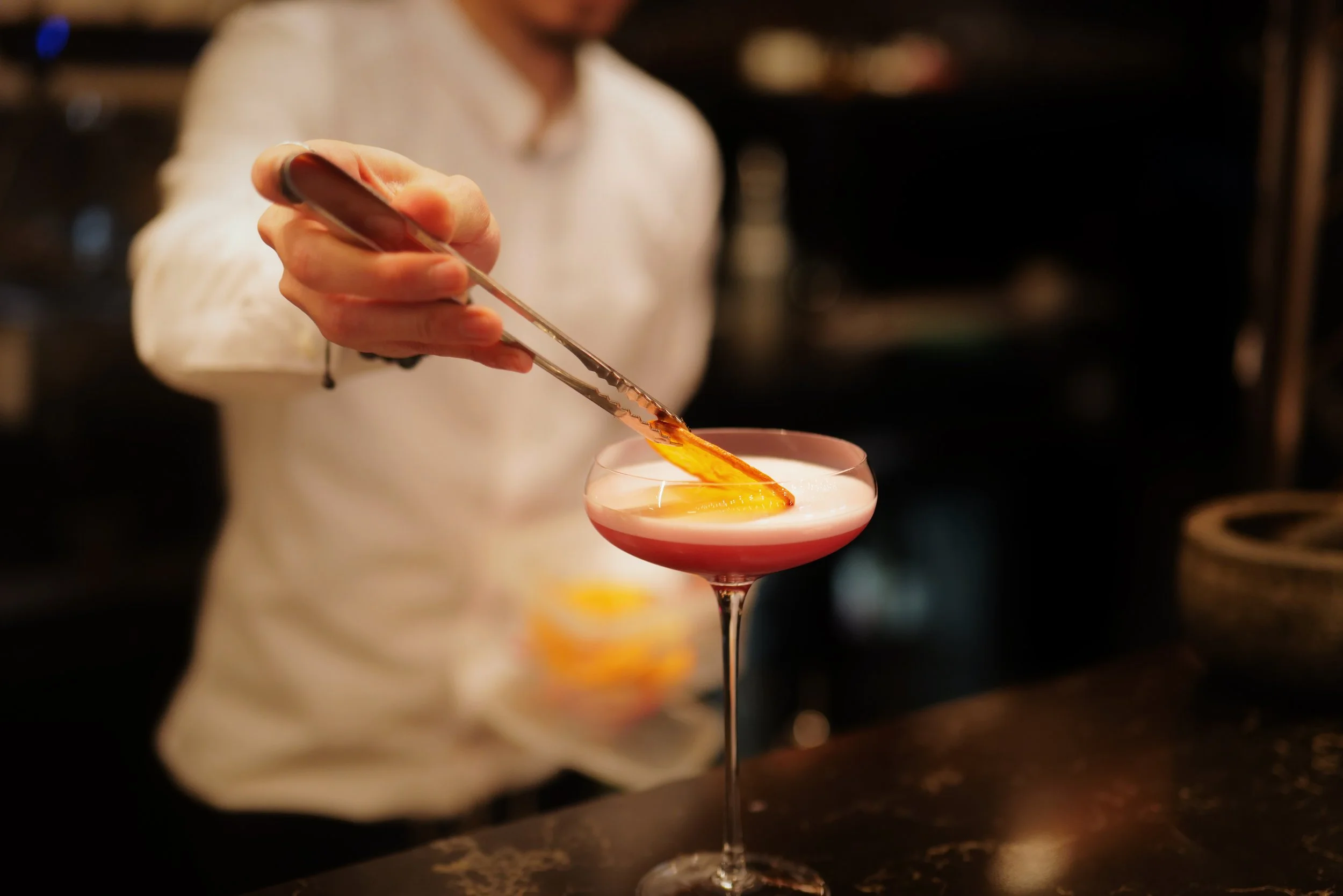 Bartender adding a garnish or ingredient to a pink cocktail in a coupe glass at a bar.