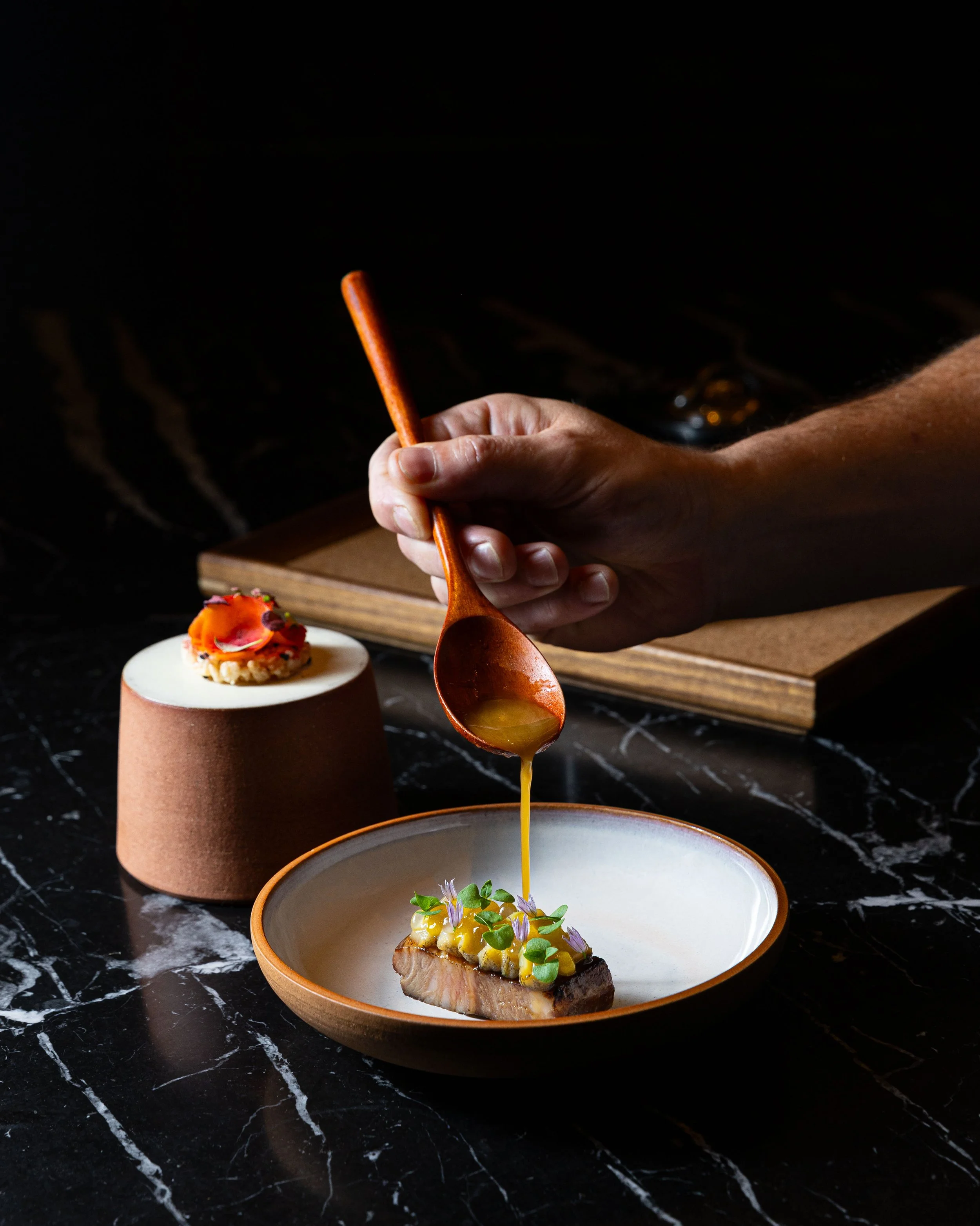 A chef pouring sauce over a plated dish with microgreens and edible flowers, with a dessert on a stand in the background on a dark marble surface.