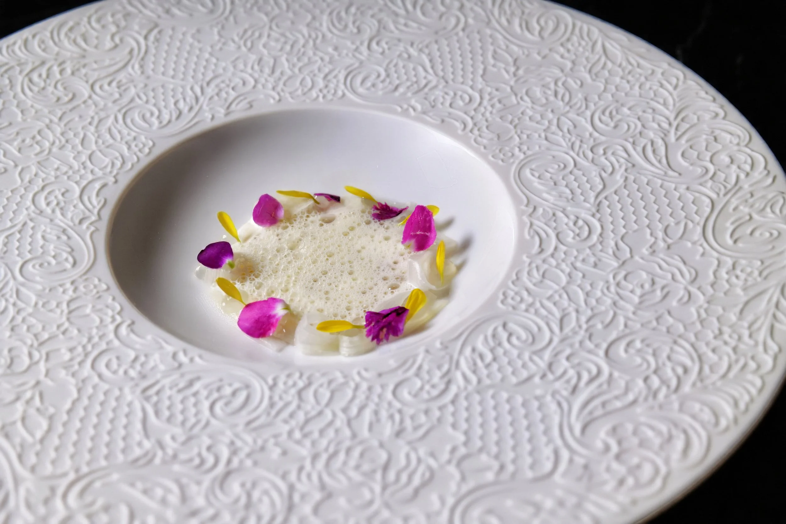 A white textured plate with an intricate embossed pattern, holding a small amount of foam or sauce garnished with pink and purple edible flowers and yellow flower petals arranged in a circle.