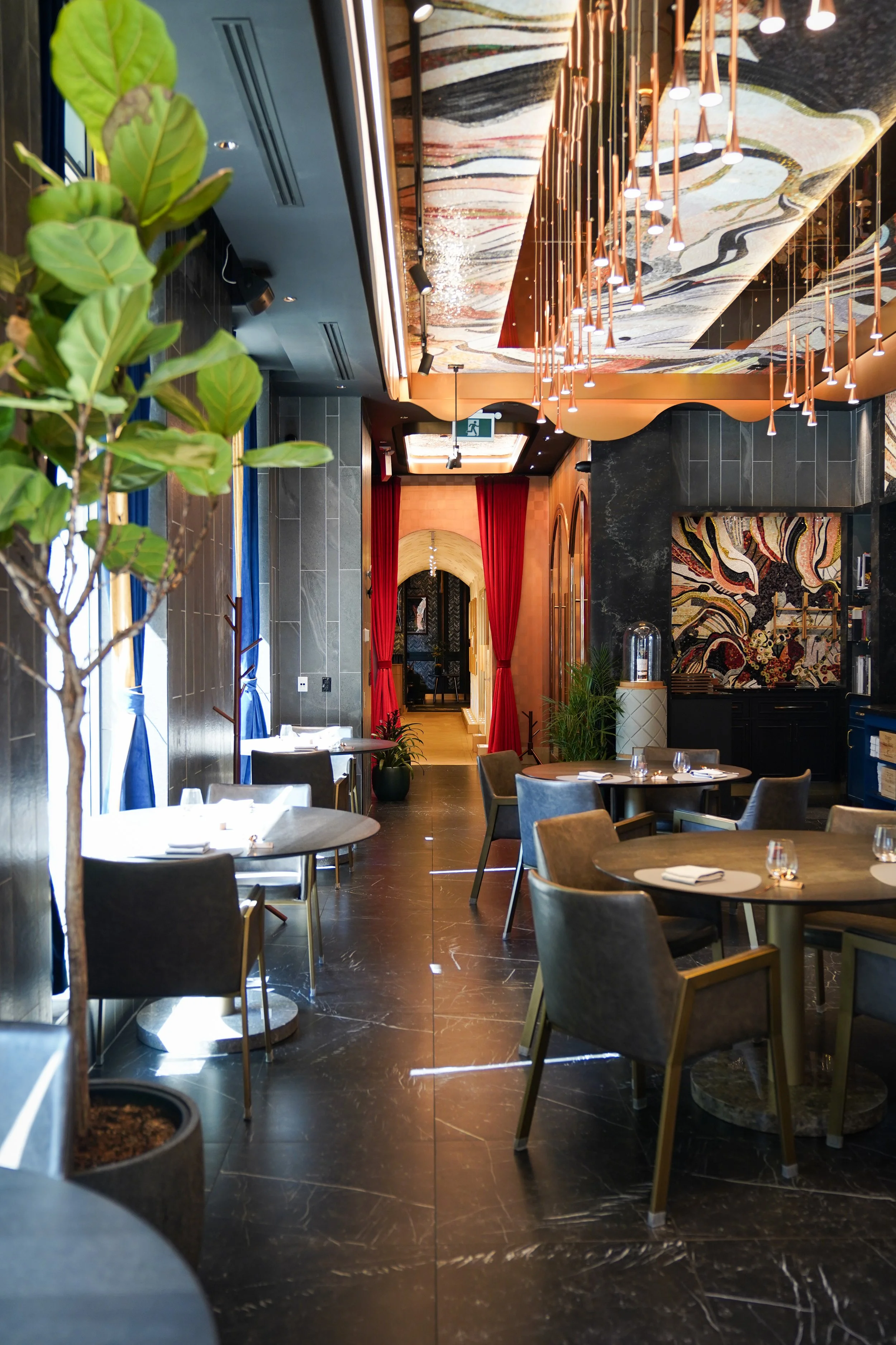 Interior of a stylish restaurant with dark marble flooring, modern tables and chairs, colorful ceiling art, and a red curtain archway in the background.
