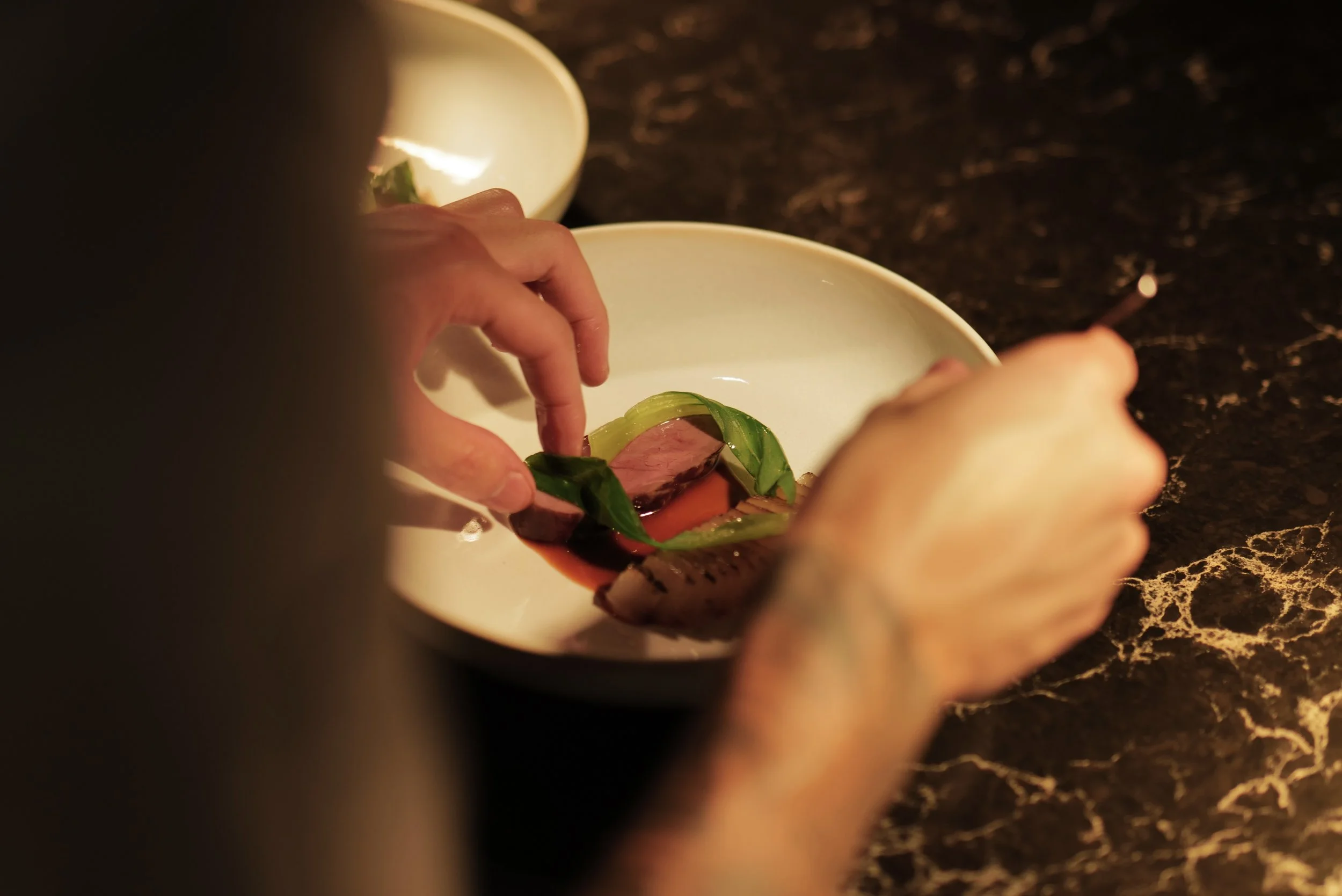 A person preparing an elegant dish with slices of meat, vegetables, and green garnish in a white bowl on a dark marble countertop.