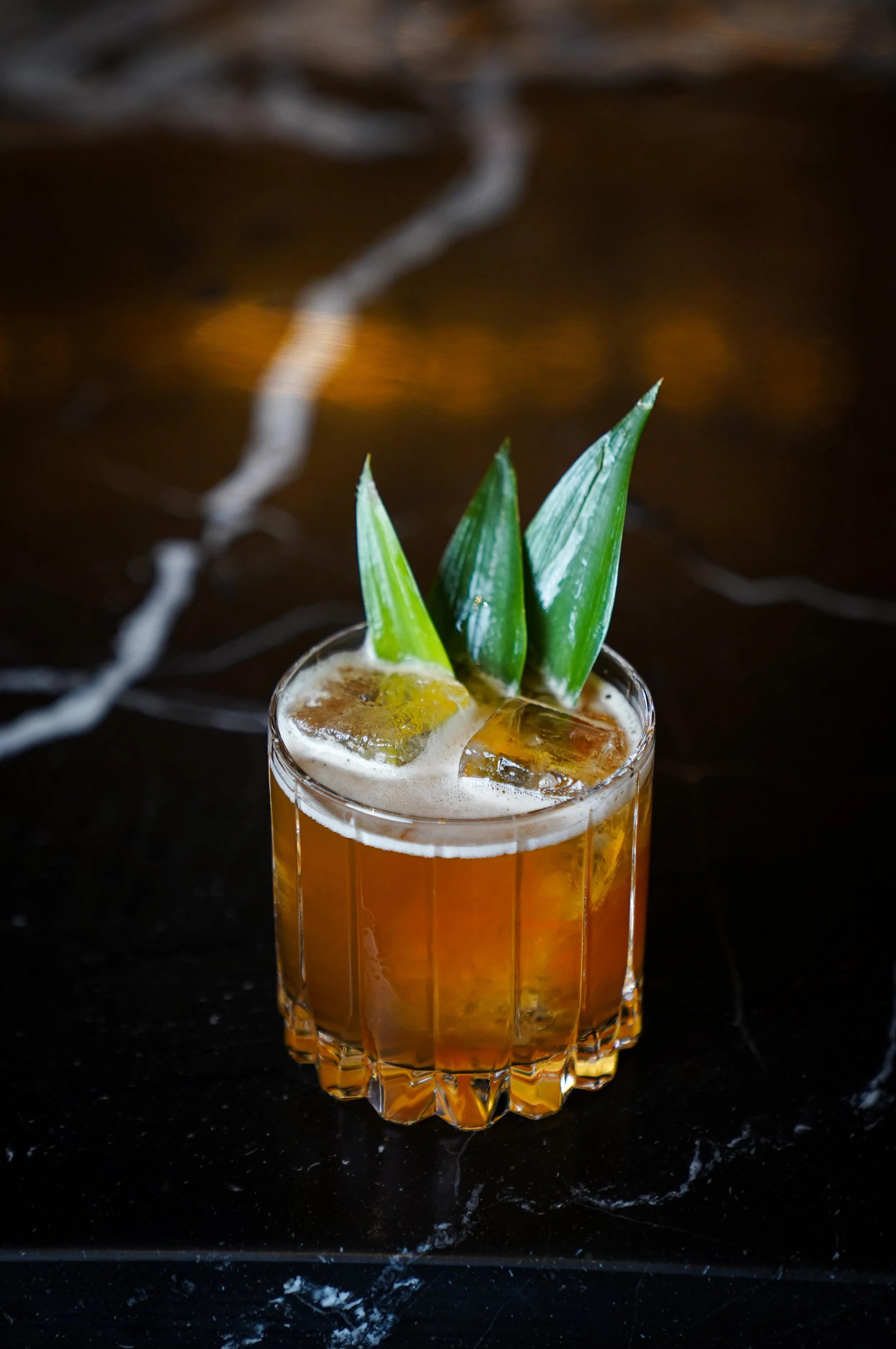 A cocktail in a clear glass garnished with pineapple leaves and ice cubes, placed on a dark marble surface.