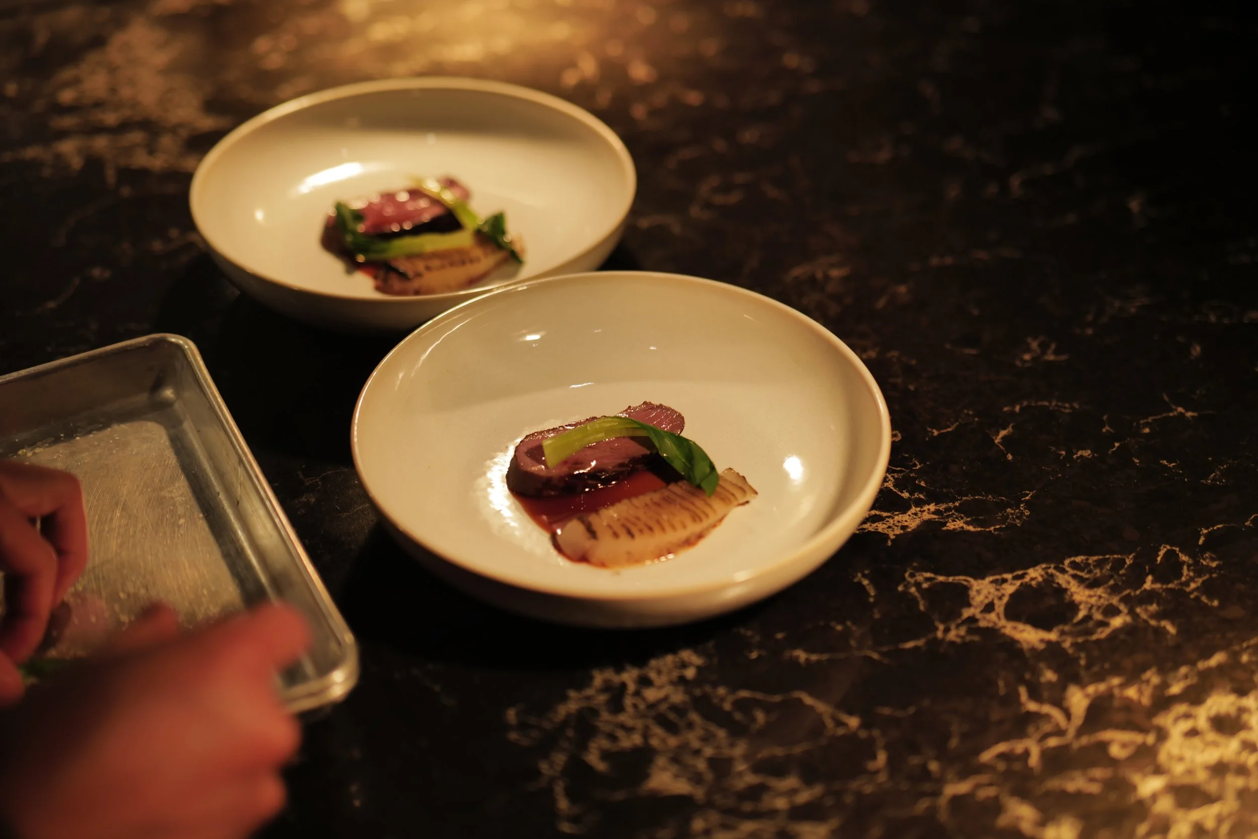 Two white ceramic bowls with slices of seared meat garnished with green herbs, placed on a dark marble countertop, with a person preparing or serving in the lower left corner.