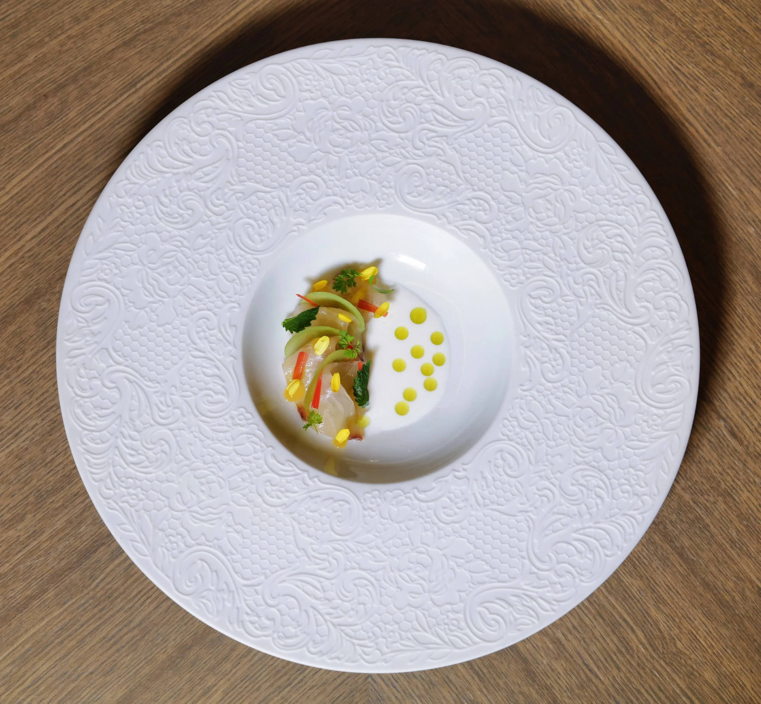 Elegant plate with a modern culinary presentation, featuring a small portion of fish topped with herbs, thin vegetable slices, and yellow garnish, surrounded by small yellow sauce dots on a decorative white plate.