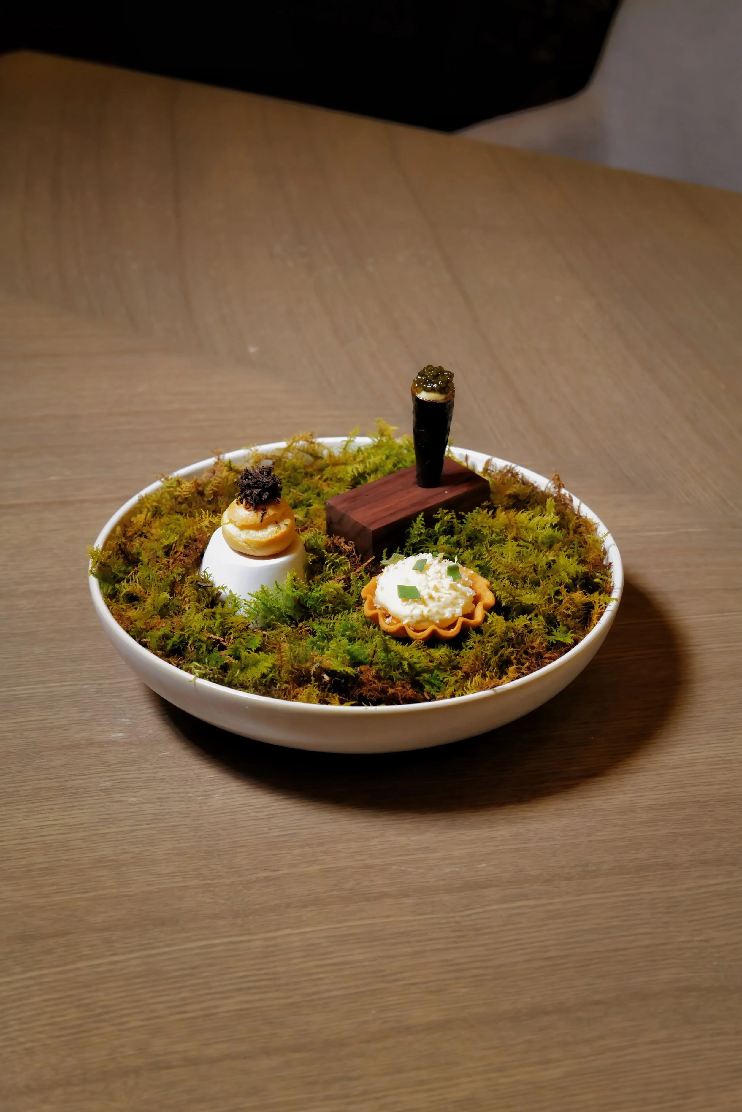 Miniature sushi and appetizers on a bed of moss in a white bowl.