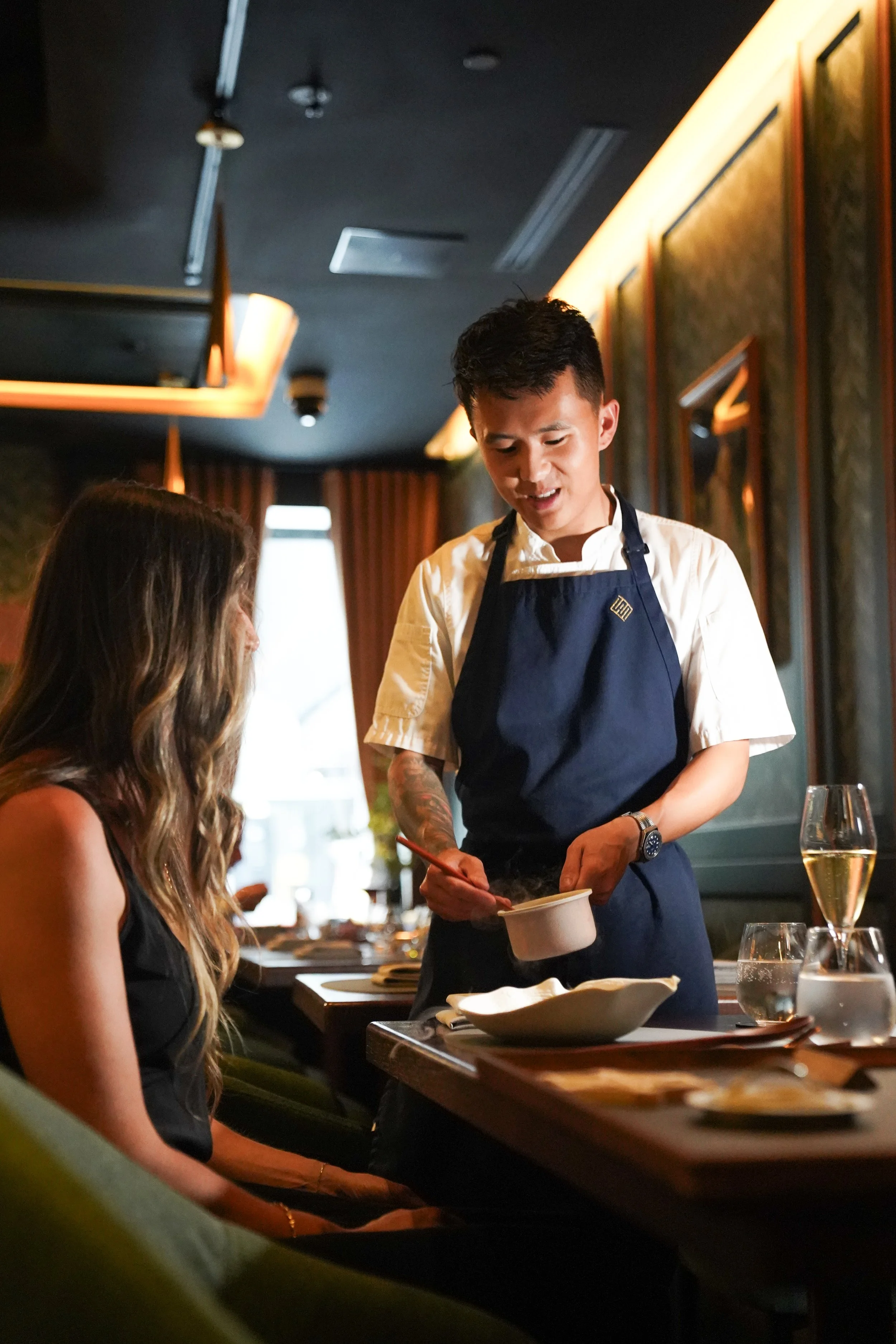 A male chef with tattoos wearing a navy apron and white shirt serves food to a female customer with long wavy hair at a restaurant table, with wine glasses on the table.