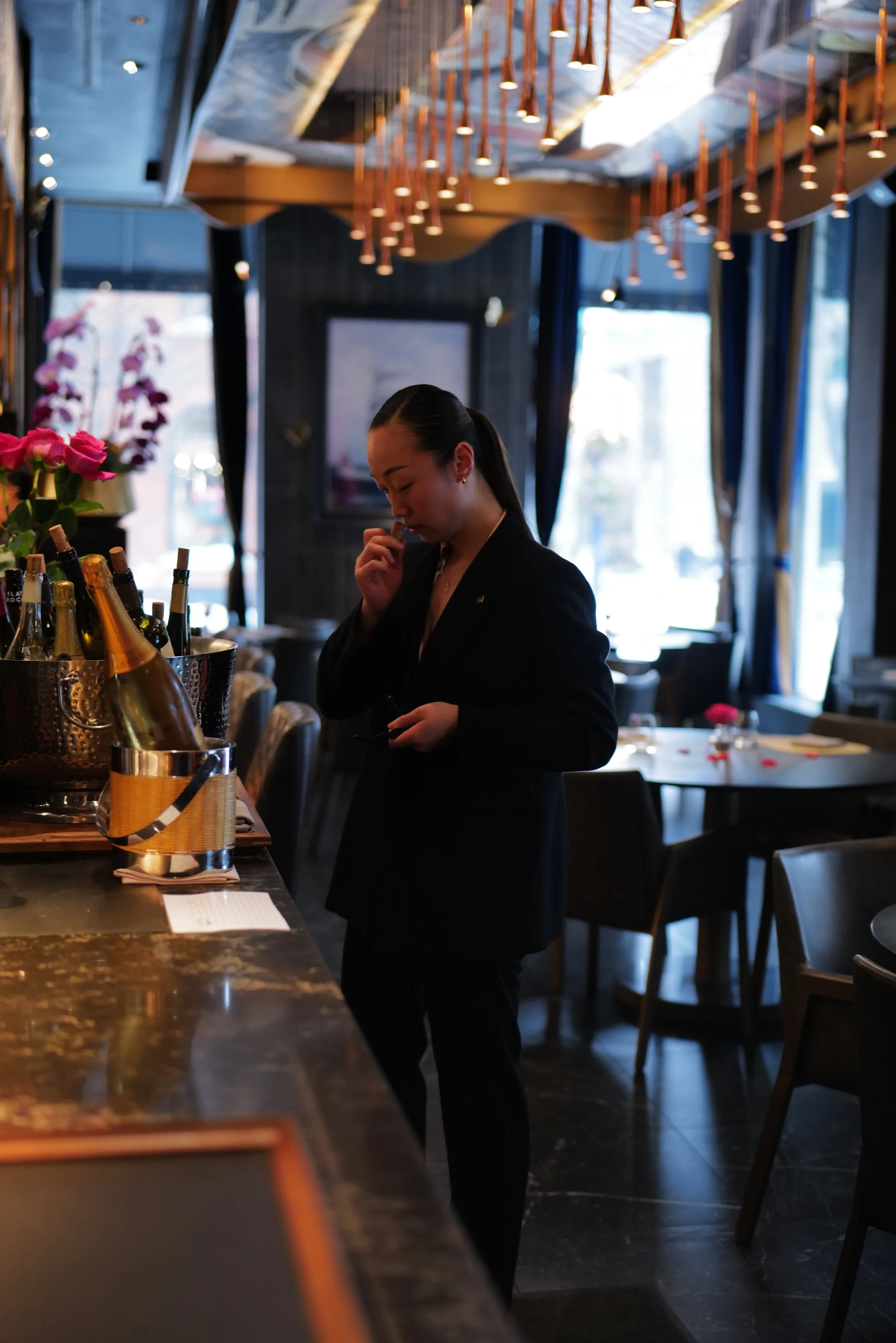 A woman dressed in a black suit standing at a bar, holding a small object near her face, with bottles of champagne and flowers on the bar counter, in a dimly lit restaurant or lounge with large windows and elegant decor.