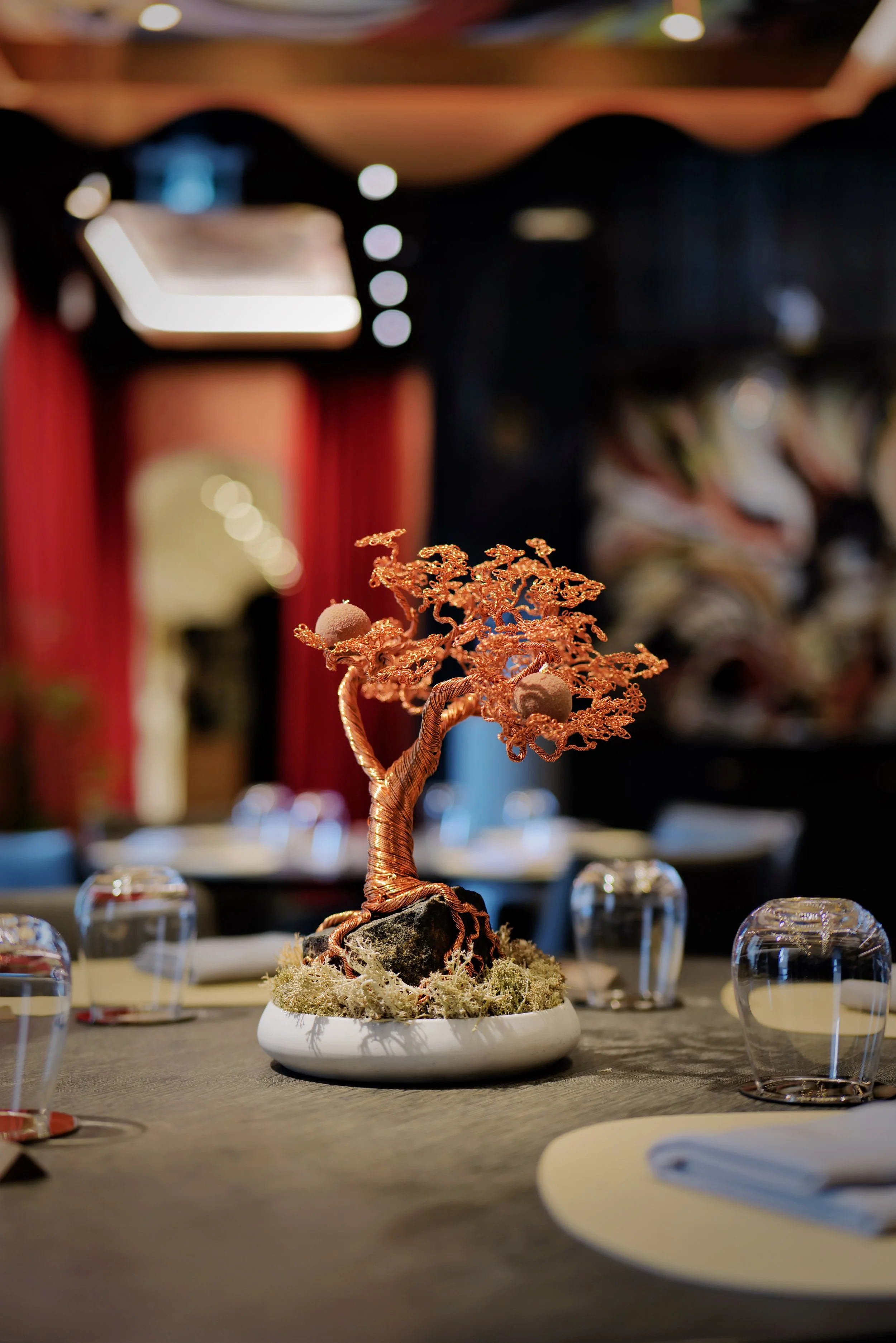 Decorative copper-colored tree sculpture on a dining table in a restaurant setting with blurred background.