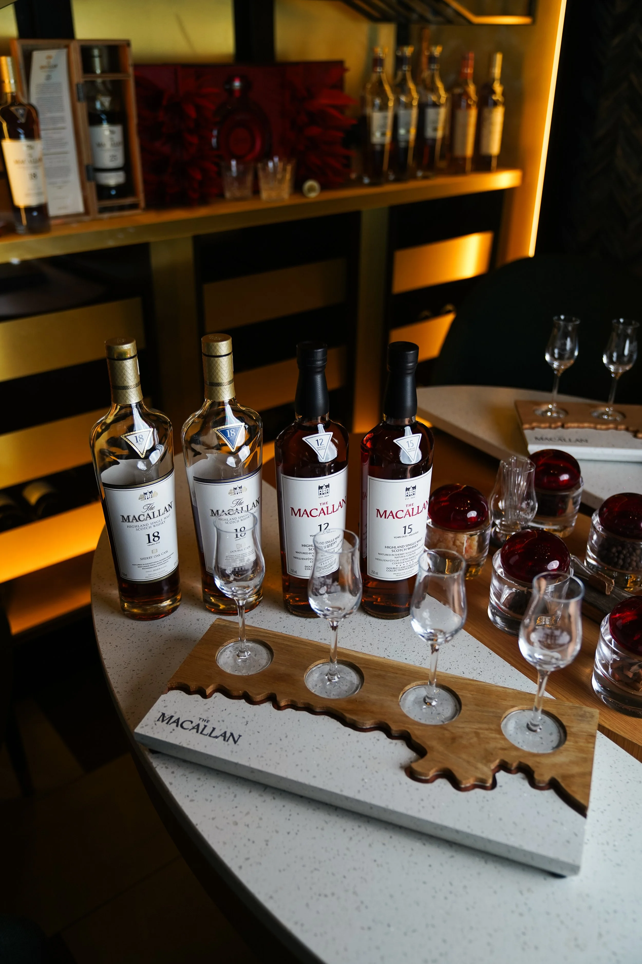 Four bottles of Macallan single malt Scotch whisky with glasses, tasting tray, and jars of cherries with chocolates on a bar counter.
