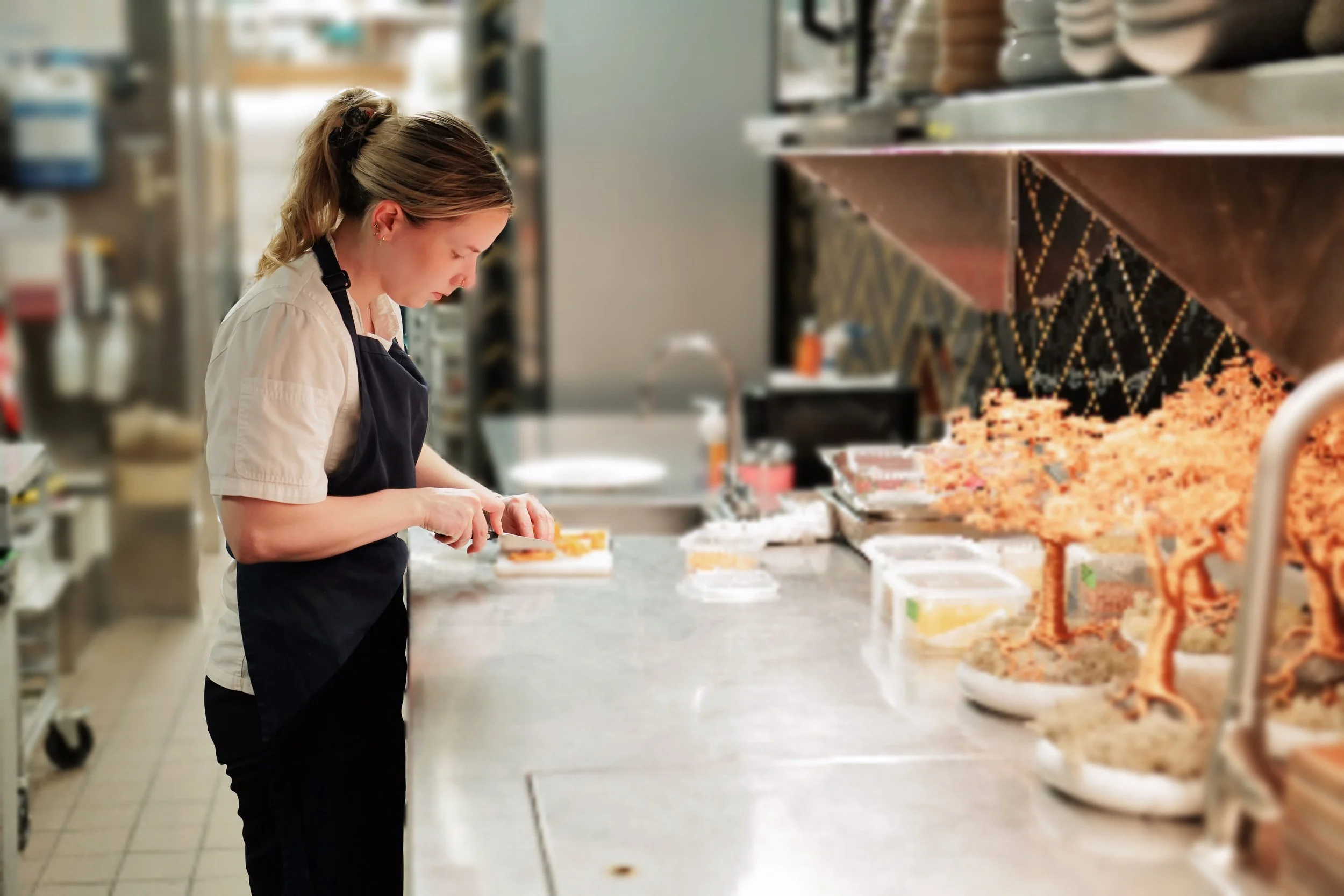 A woman in a black apron and beige shirt slicing food in a kitchen with various ingredients and kitchenware on the counter.