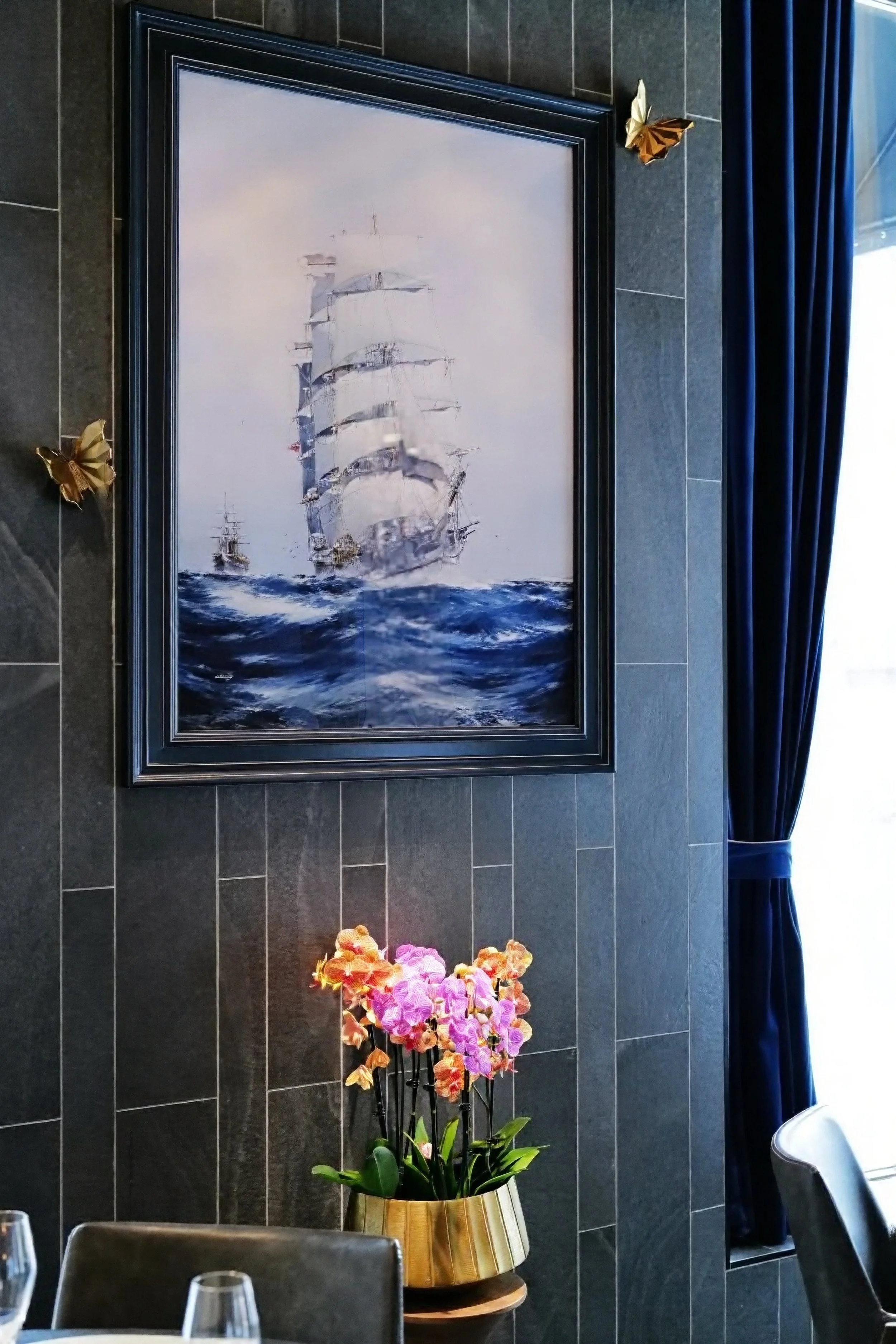A painting of a tall ship sailing on rough waters hangs on a dark tiled wall. Gold butterfly decorations are on the wall, and a pink and orange orchid plant in a gold striped pot is on a table below, next to chairs in a restaurant or cafe.