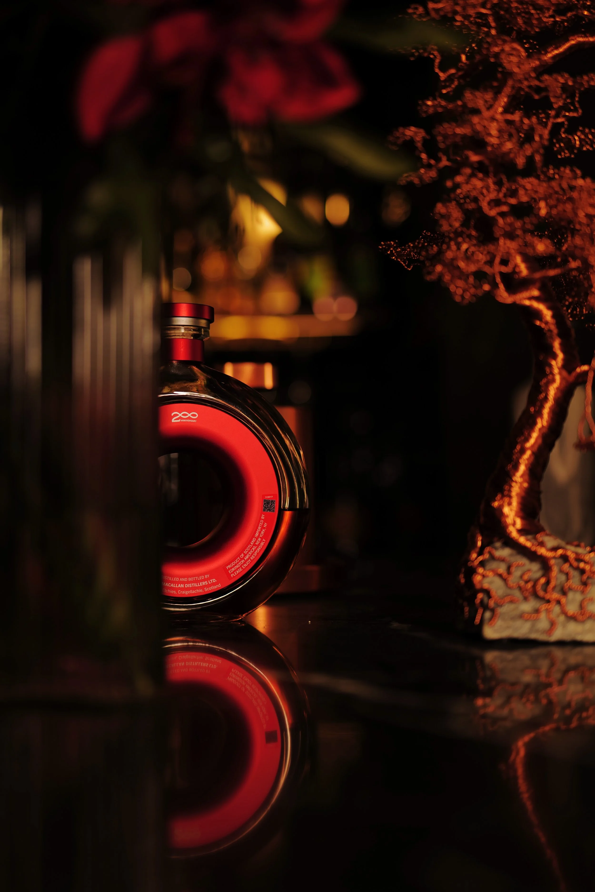 A circular bottle with a red label and black cap, placed on a dark reflective surface, surrounded by blurred floral decorations and a decorative tree with orange lights.