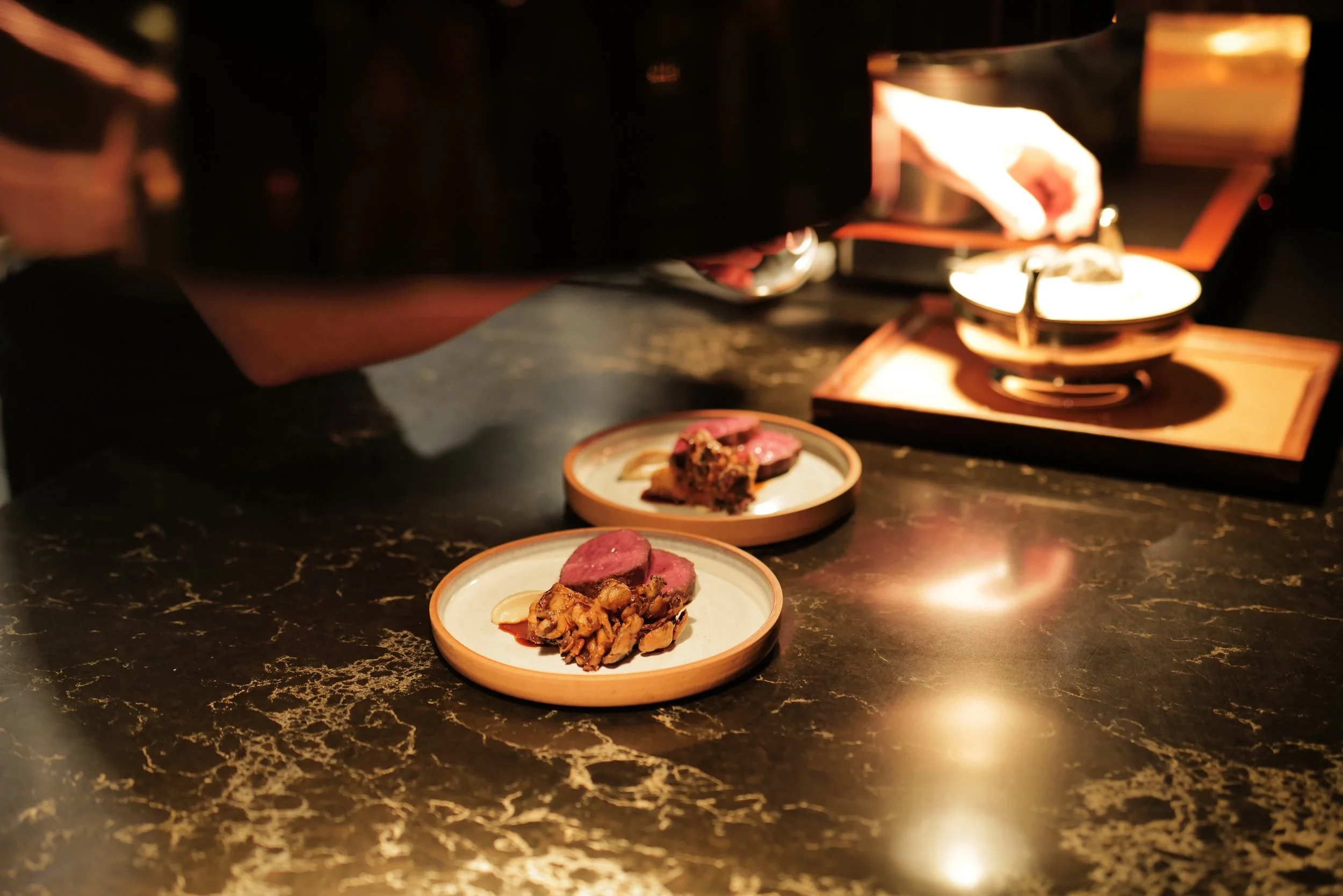 Two small plates with slices of cooked meat and garnishes on a black marble countertop, with a person serving food from a warming tray in the background.
